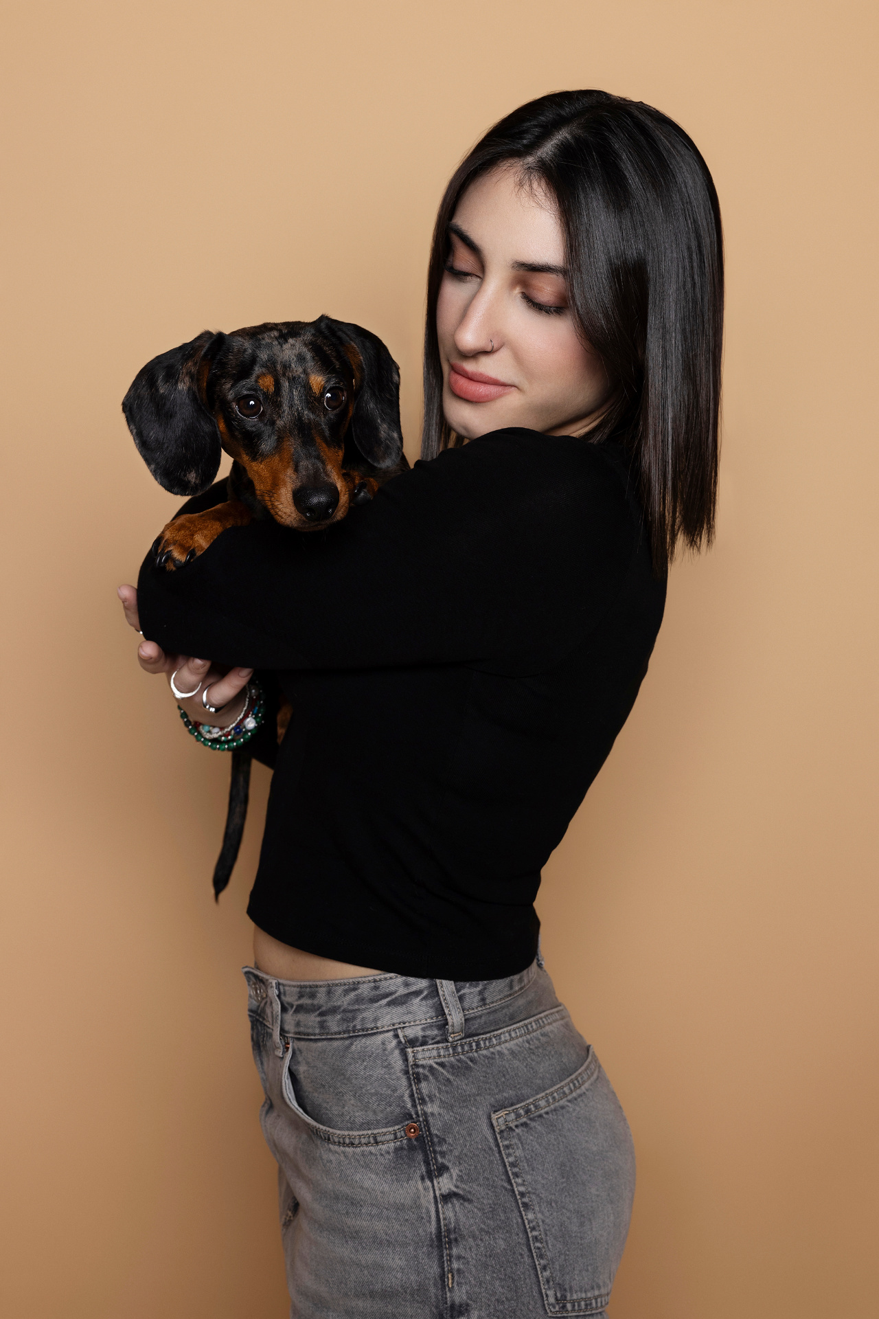 Donna con capelli scuri abbraccia affettuosamente un bassotto nero e marrone su sfondo beige. Ritratto fine art che celebra il legame tra umano e animale, scattato da Claudia Spaziani a Bologna.