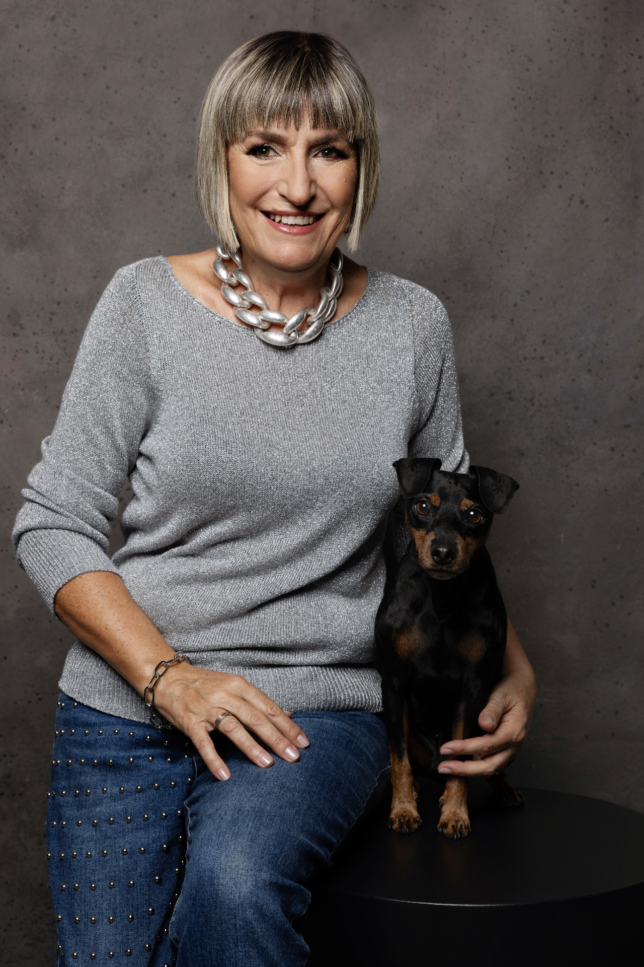 Donna con capelli grigio-biondi e sorriso tenero accanto a Miniature Pinscher nero focato, in posa su superficie nera. Fotografia fine art di Claudia Spaziani, Bologna.