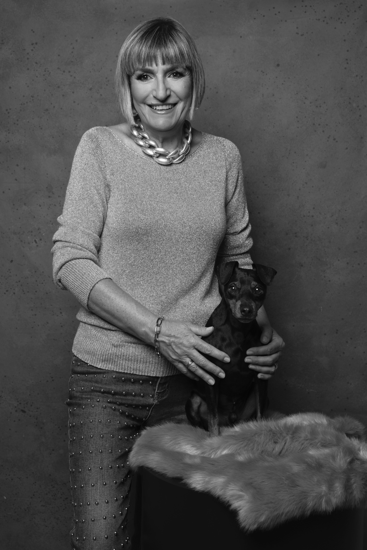 Ritratto in bianco e nero di donna sorridente accanto a Miniature Pinscher seduto su superficie pelosa, gesto affettuoso. Fotografia fine art di Claudia Spaziani, Bologna.