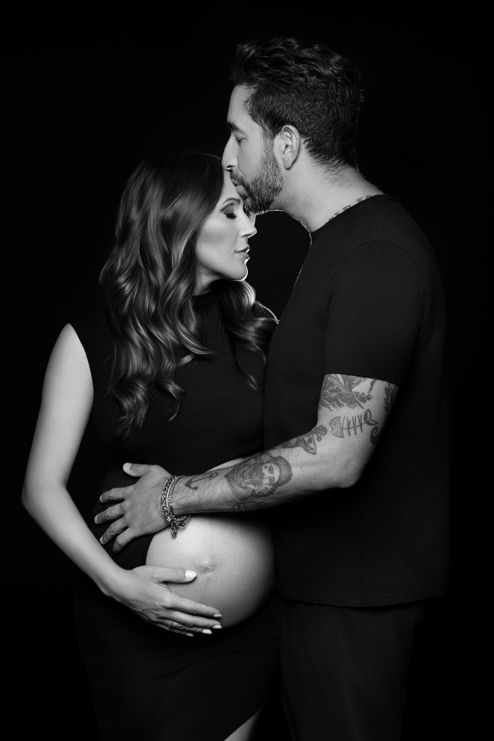 Fotografia in bianco e nero su fondo scuro: lui le bacia la fronte mentre entrambi tengono le mani sul pancione scoperto. Studio fotografico di maternità a Bologna.