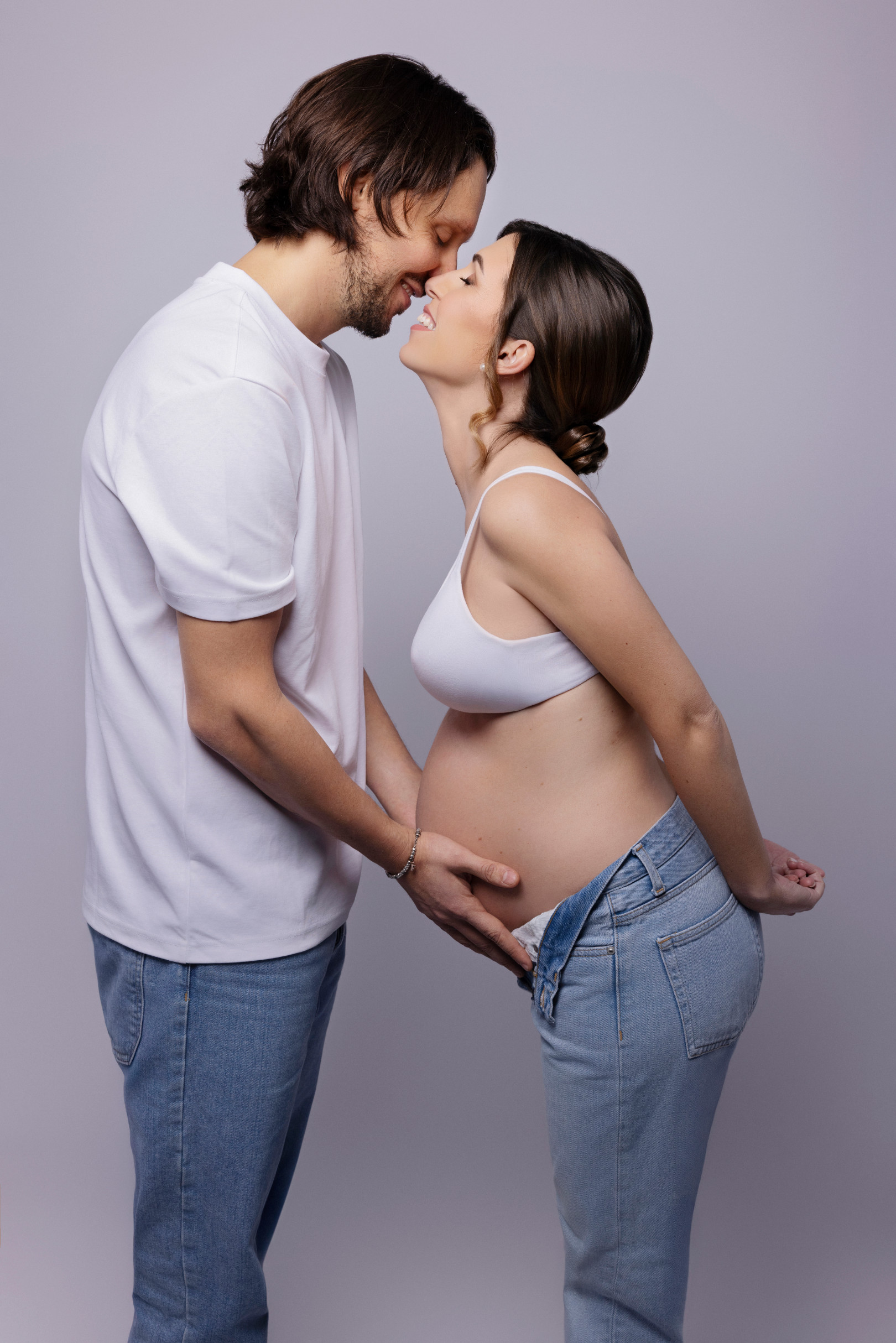 Coppia in jeans e t-shirt bianca si sfiora con il naso mentre lui appoggia le mani sul pancione scoperto. Fotografia di gravidanza in studio, Bologna.