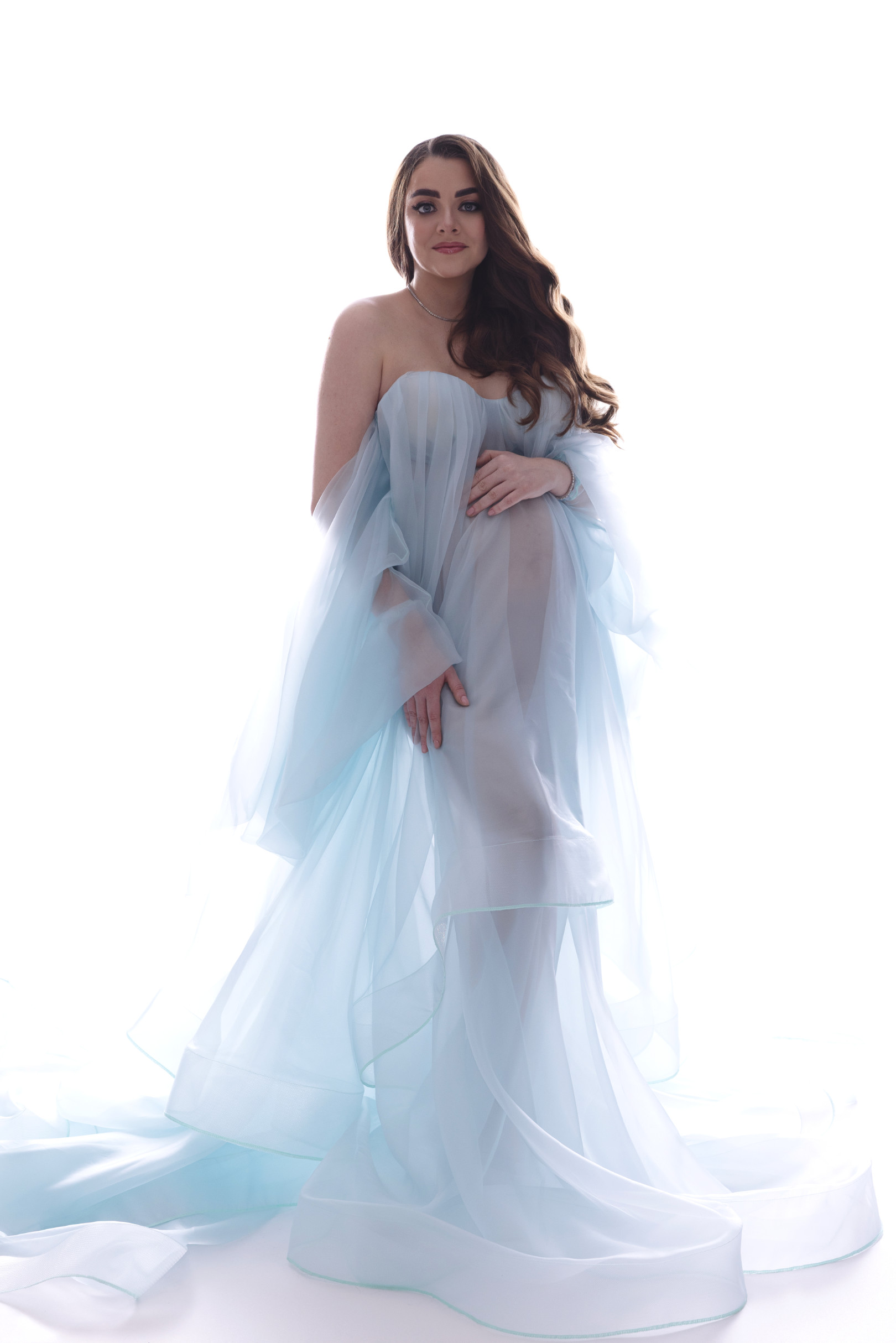 Donna incinta in abito di organza azzurra a strati con ampio strascico, in piedi su fondo bianco luminoso. Ritratto di maternità realizzato a Bologna da Claudia Spaziani.