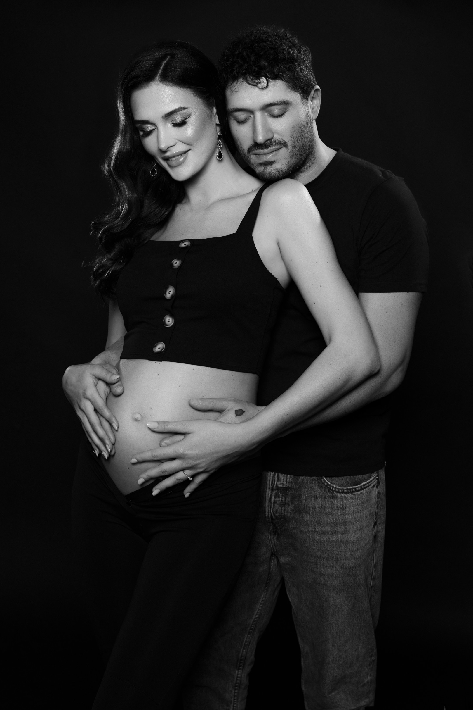Ritratto di coppia in bianco e nero su fondo scuro: futura mamma in top nero abbottonato, il compagno alle spalle con le mani sul pancione. Fotografia di Claudia Spaziani, Bologna.