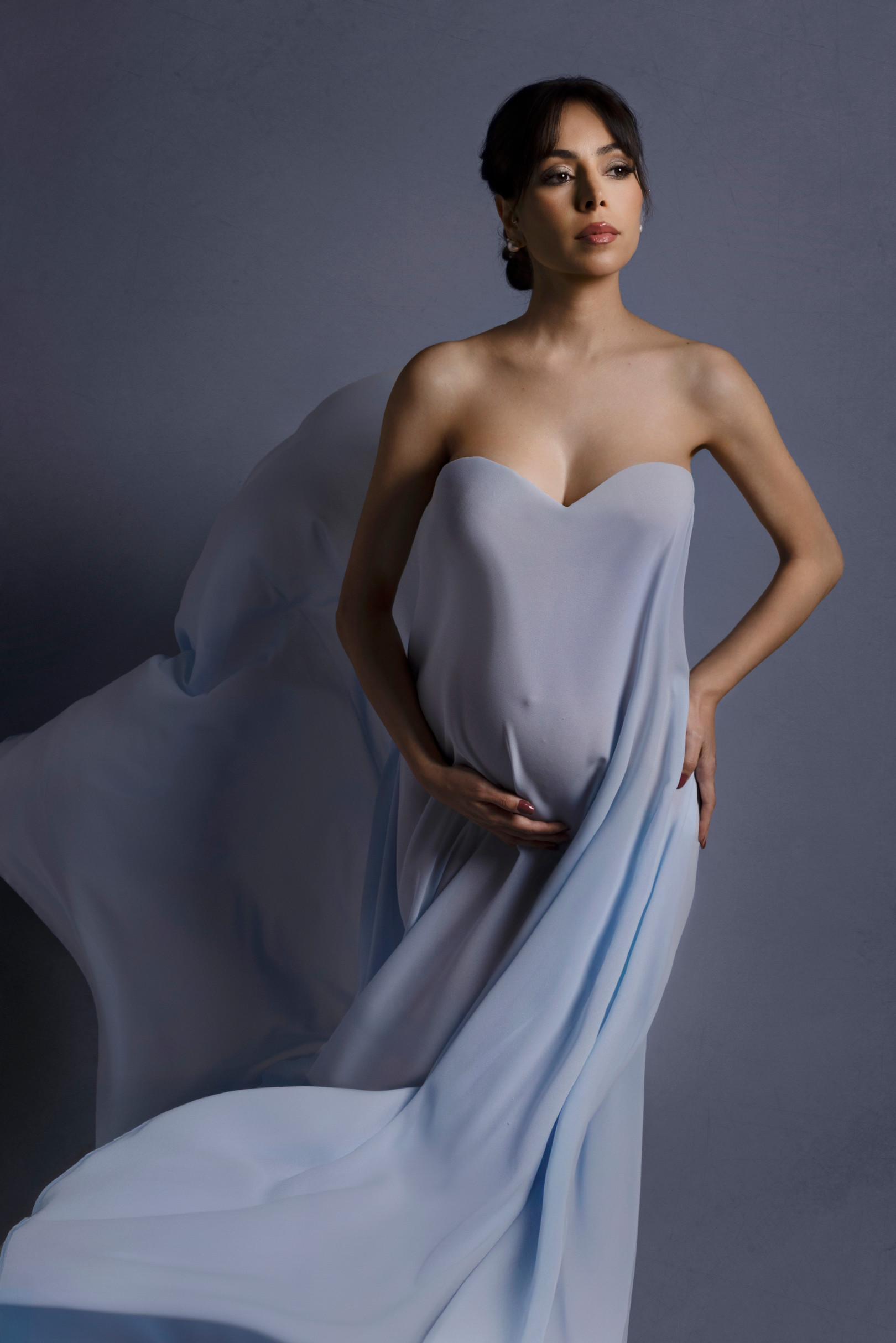 Ritratto premaman in abito azzurro polvere con corpetto a cuore e lungo velo di chiffon in movimento, mano sul pancione. Studio fotografico Claudia Spaziani, Bologna.