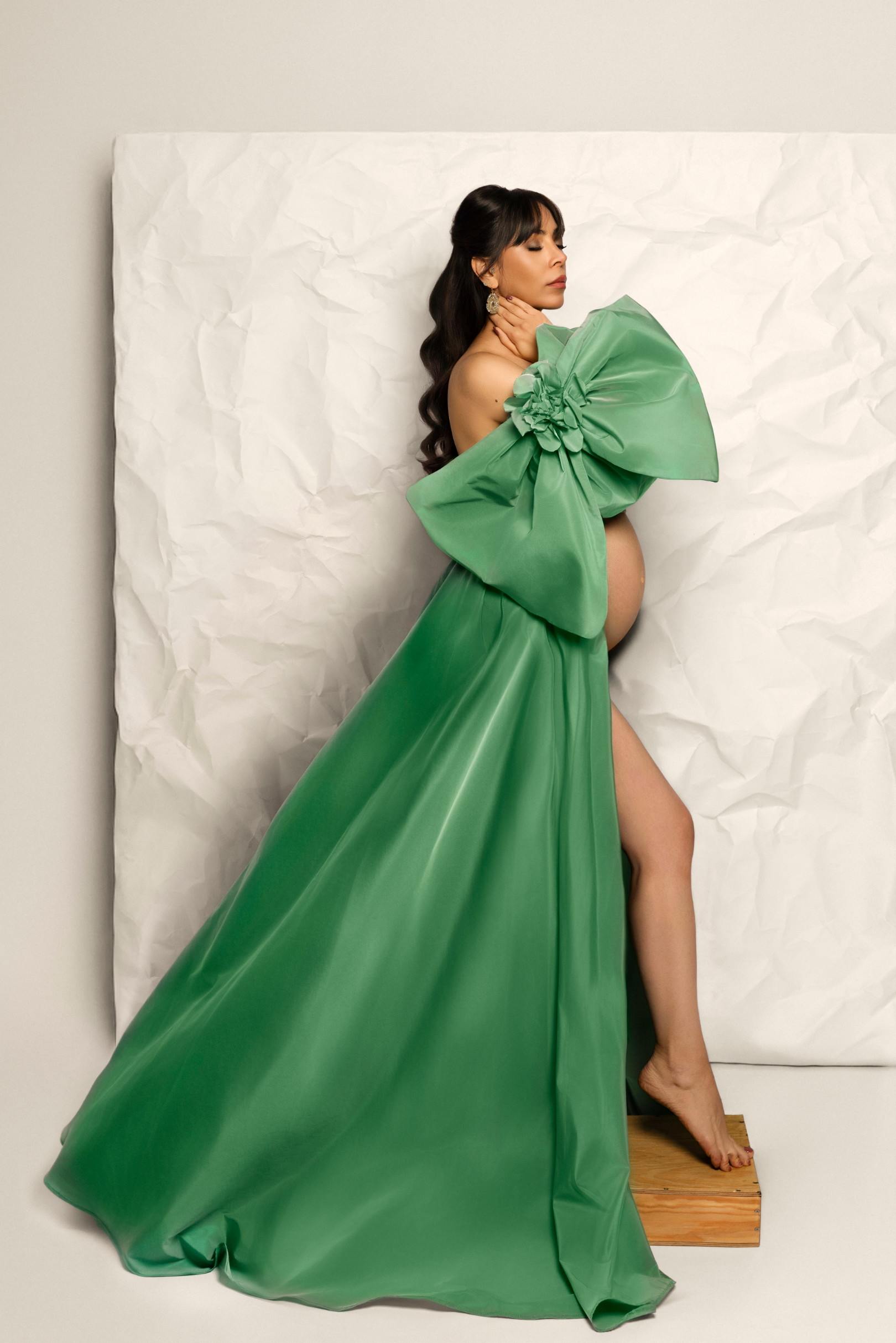 Donna incinta di profilo in abito di raso verde con enorme fiocco floreale e spacco profondo, davanti a un fondale di carta stropicciata. Claudia Spaziani, Bologna.