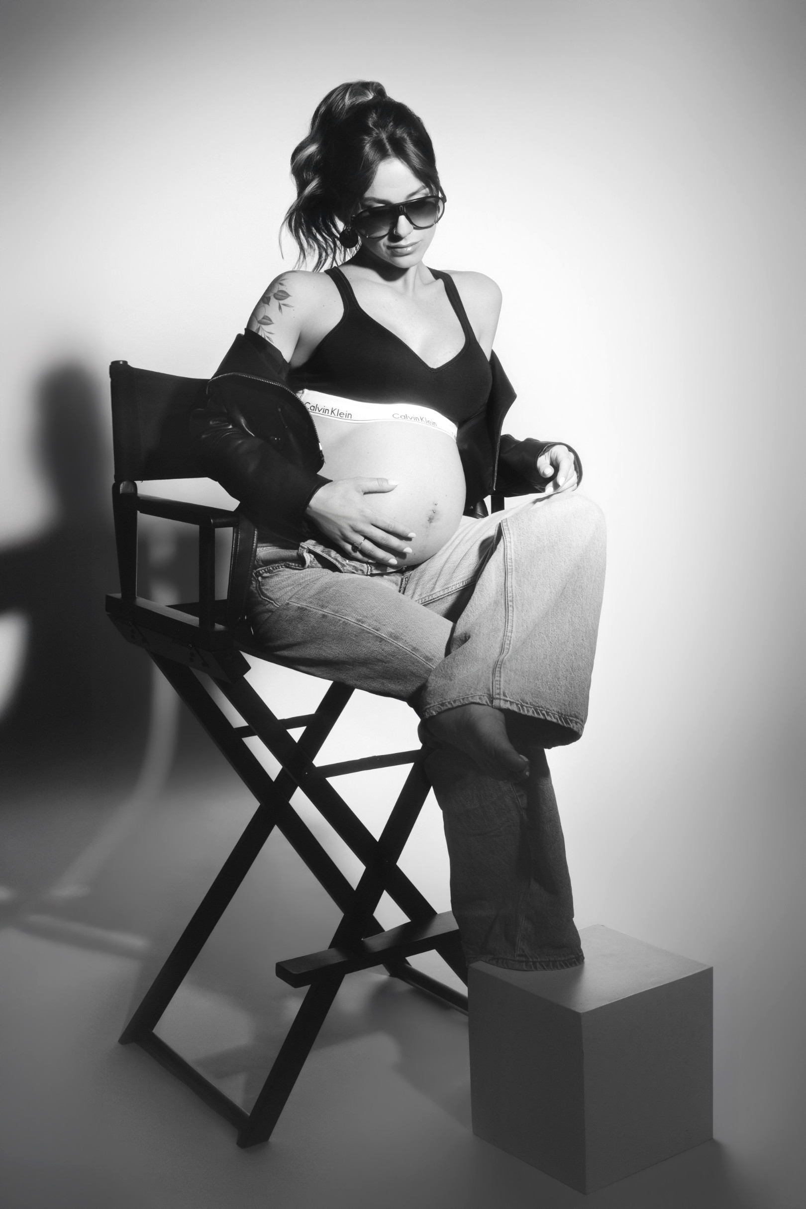Ritratto in bianco e nero: donna incinta seduta su una sedia da regista con occhiali da sole, giacca di pelle e jeans sbottonati. Studio fotografico Claudia Spaziani.