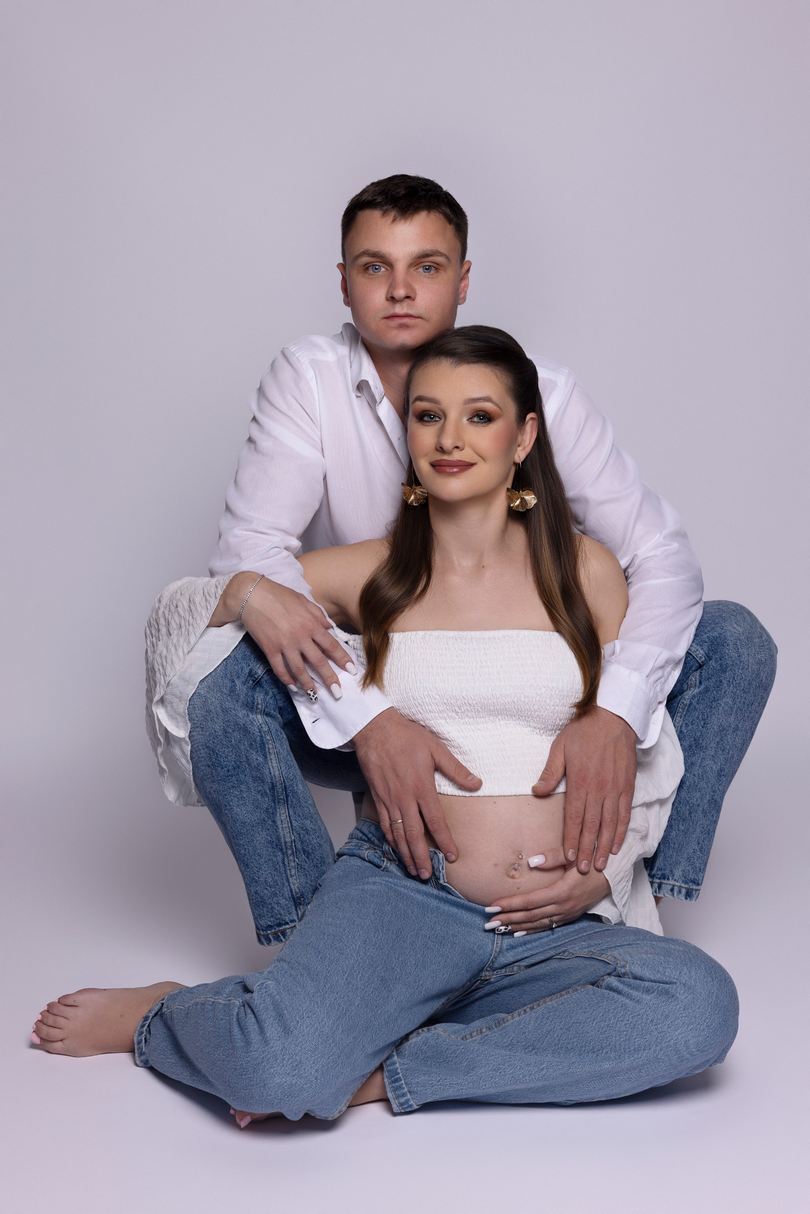 Coppia in attesa seduta a terra su fondo grigio chiaro, entrambi in camicia bianca e jeans, mani di lui sul pancione. Studio fotografico Claudia Spaziani, Bologna.