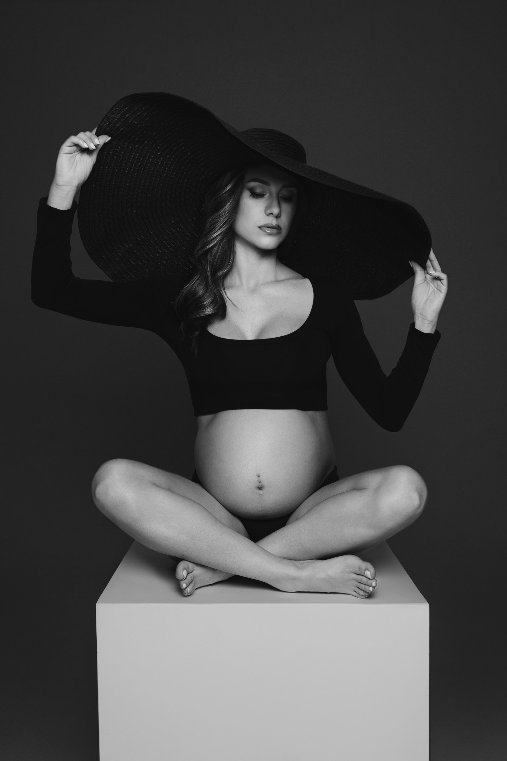 Ritratto in bianco e nero: donna incinta seduta a gambe incrociate su un cubo bianco, top nero corto e cappello a tesa larghissima. Claudia Spaziani, Bologna.