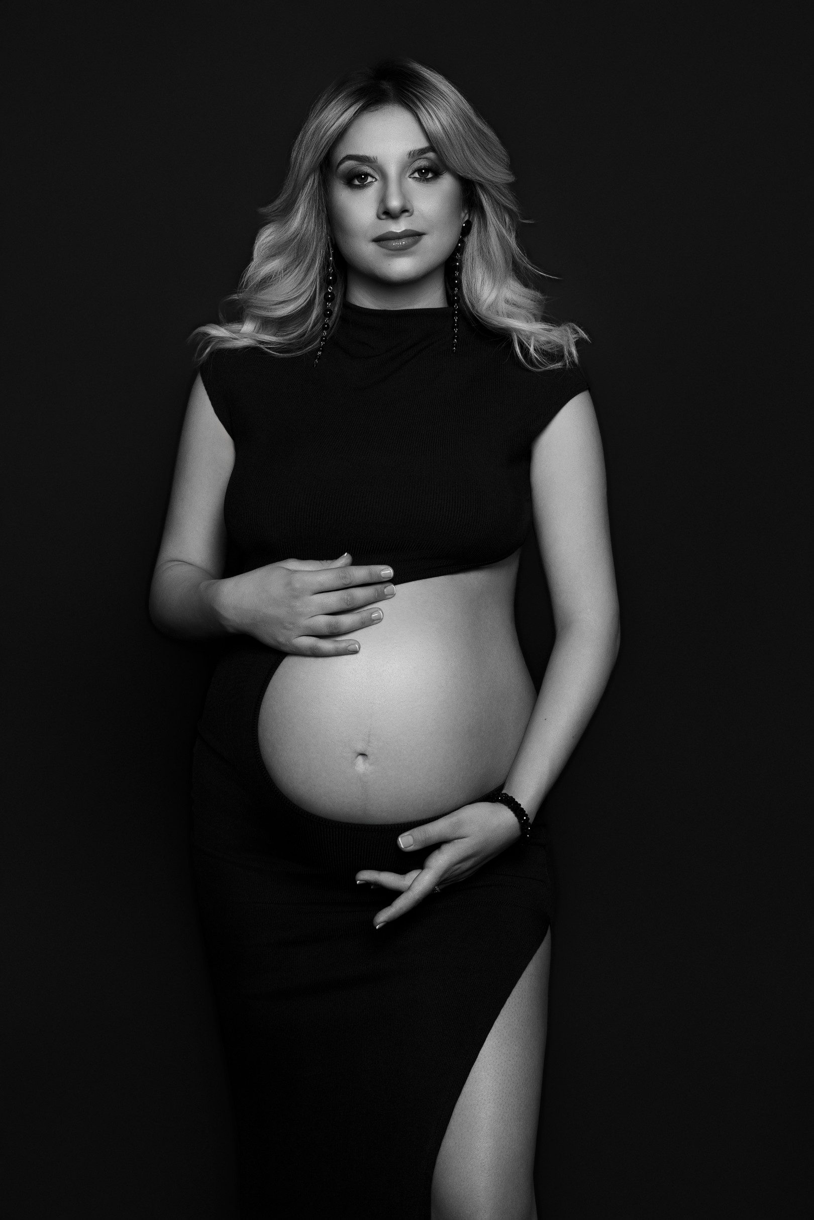 Ritratto in bianco e nero su fondo scuro: donna incinta in completo nero con apertura sul pancione e orecchini pendenti. Servizio maternità in studio a Bologna.