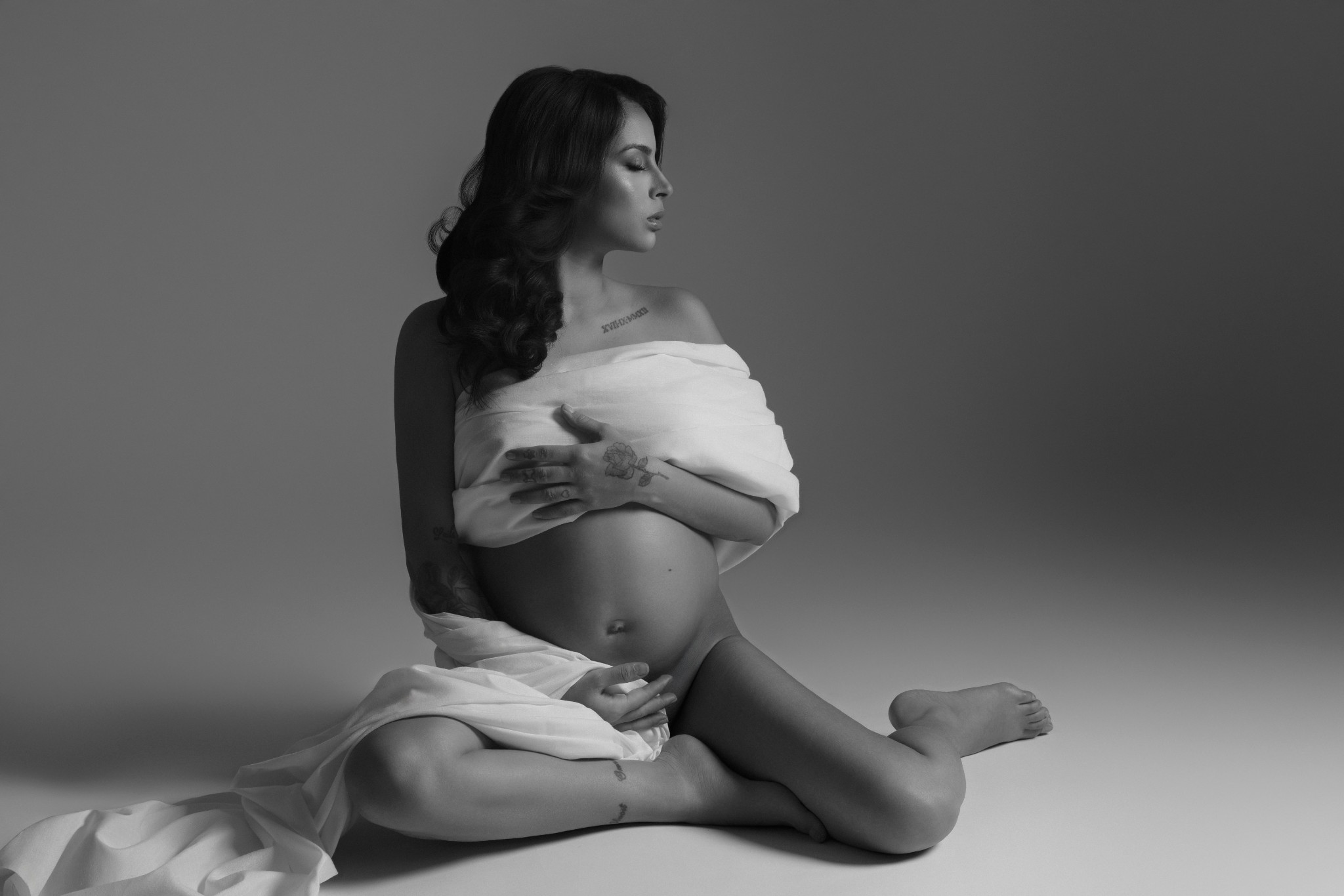 Donna incinta seduta a terra avvolta in un drappo bianco sceso sulle spalle, pancione scoperto, in bianco e nero. Studio fotografico Claudia Spaziani, Bologna.