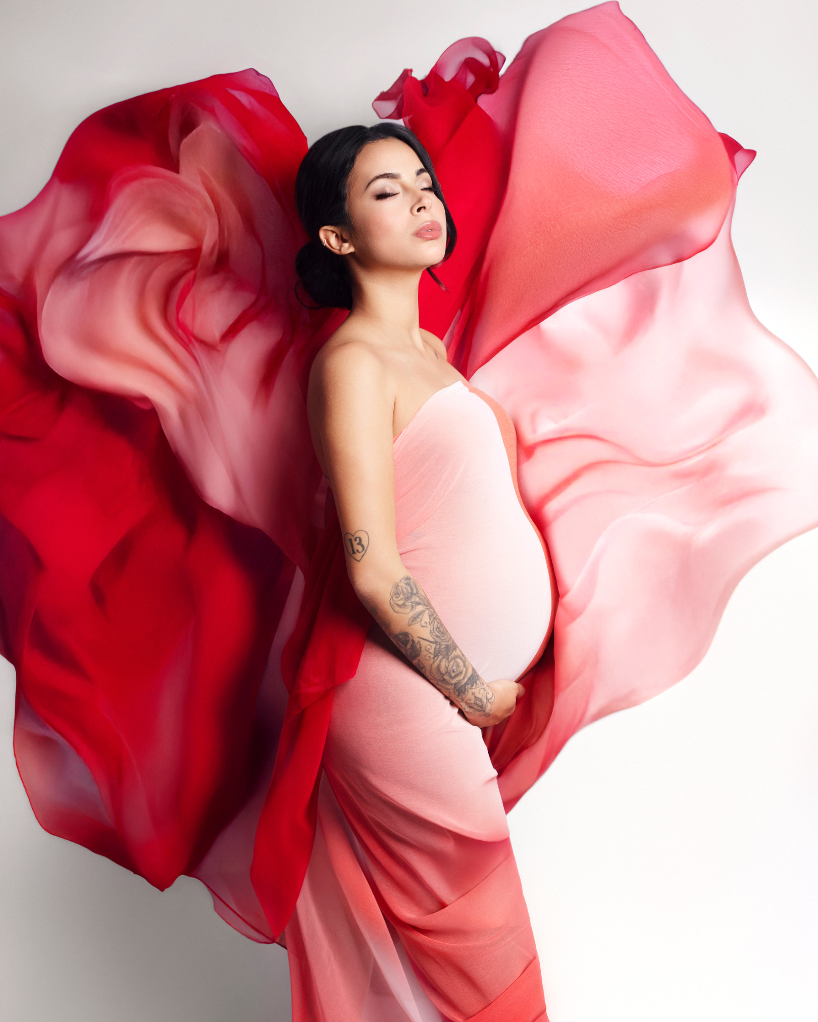 Donna incinta in abito rosa sfumato con ampi teli di chiffon rosso sospesi in aria alle sue spalle. Servizio fotografico premaman di Claudia Spaziani, Bologna.