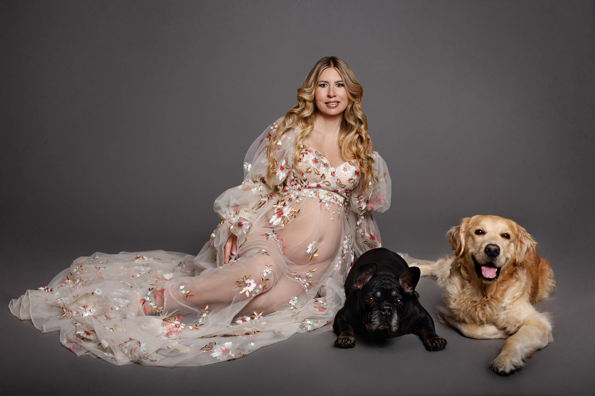 Futura mamma seduta a terra in abito di tulle ricamato a fiori, accompagnata da un bulldog francese nero e un golden retriever. Fotografia di Claudia Spaziani, Bologna.