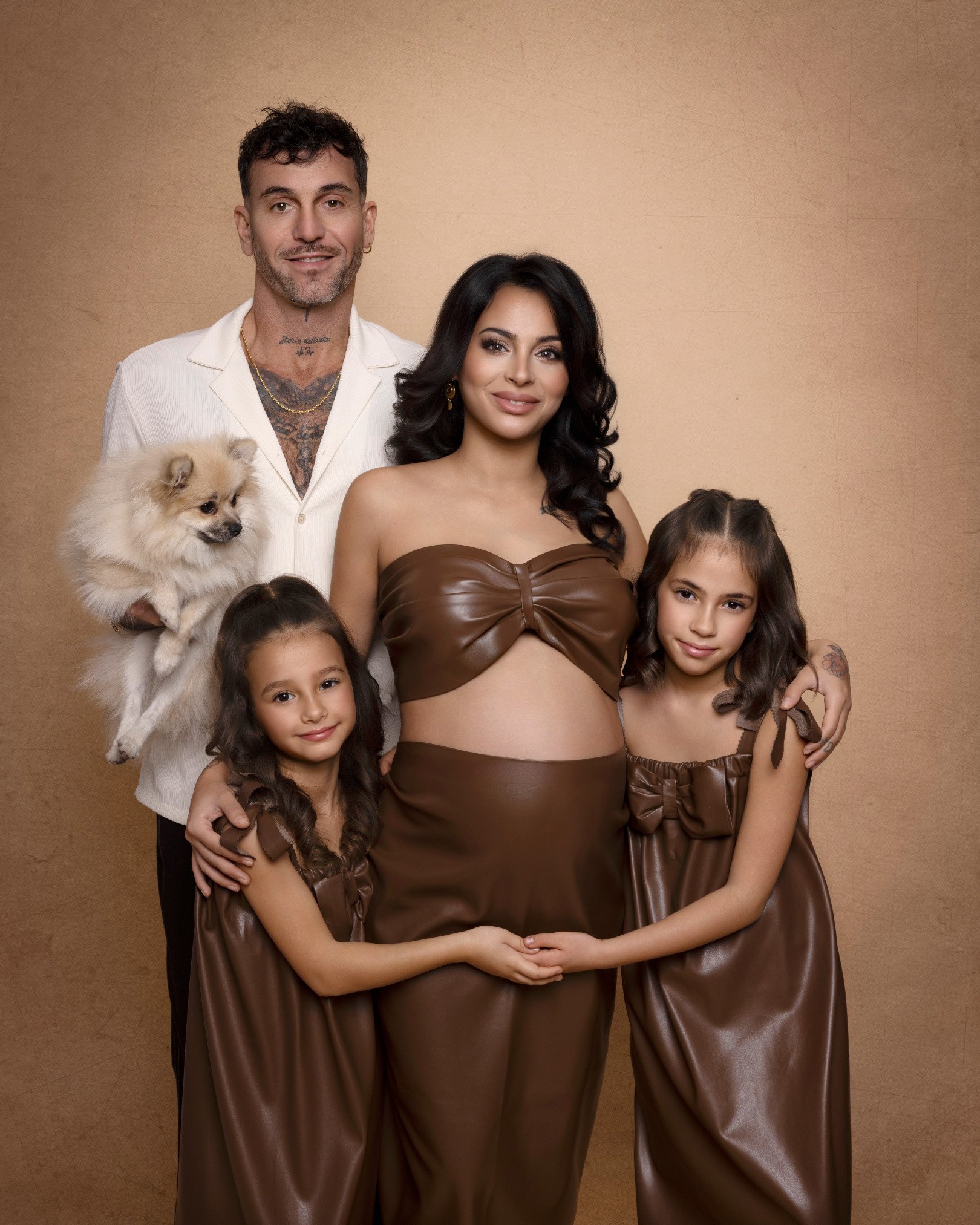 Ritratto di famiglia su fondale beige: futura mamma in completo marrone, il compagno con un cane pomerania in braccio e due bambine in abiti coordinati. Claudia Spaziani.