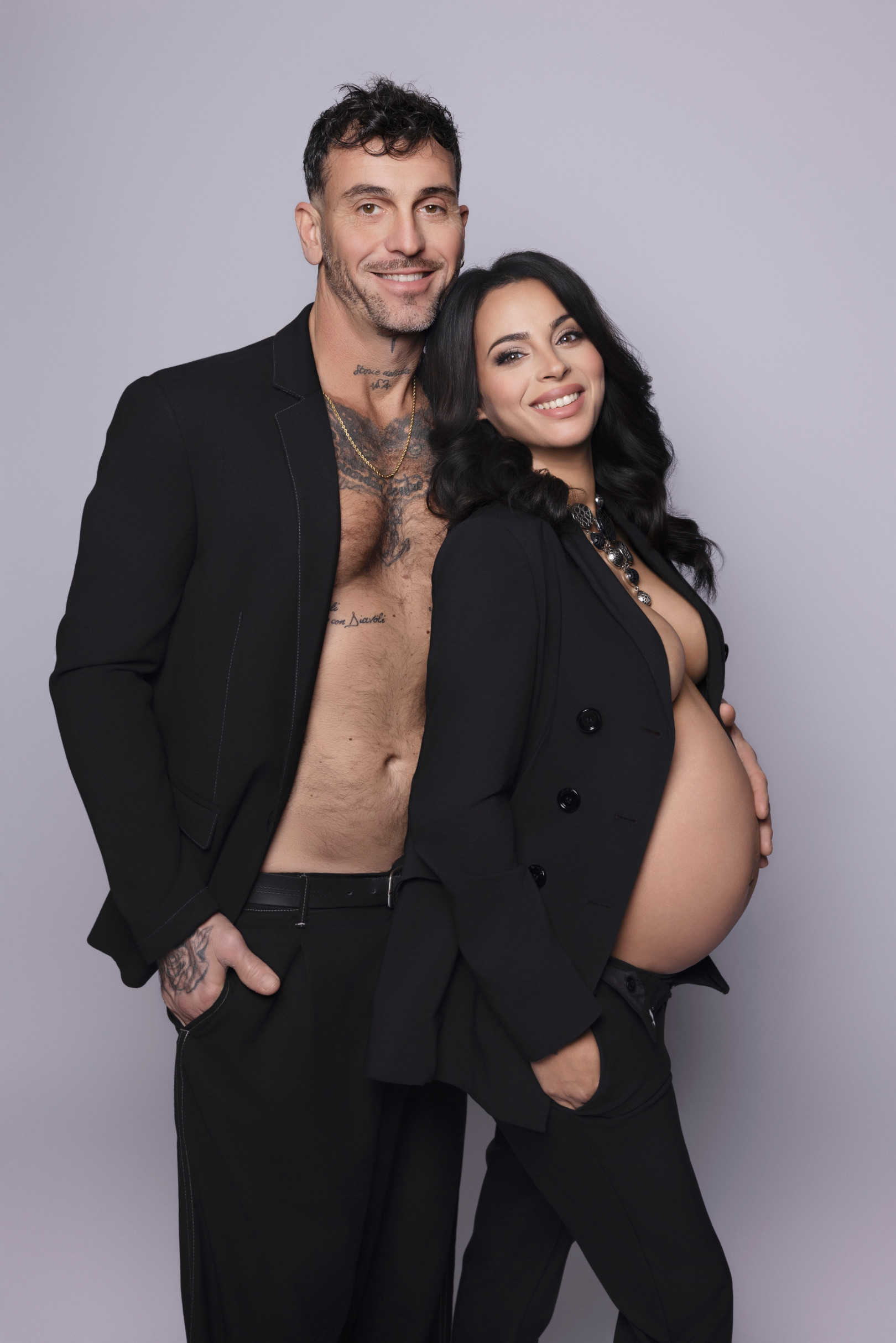 Coppia in completo nero su fondo lilla: lei con la giacca aperta sul pancione, lui con blazer indossato a pelle. Studio fotografico di maternità a Bologna.