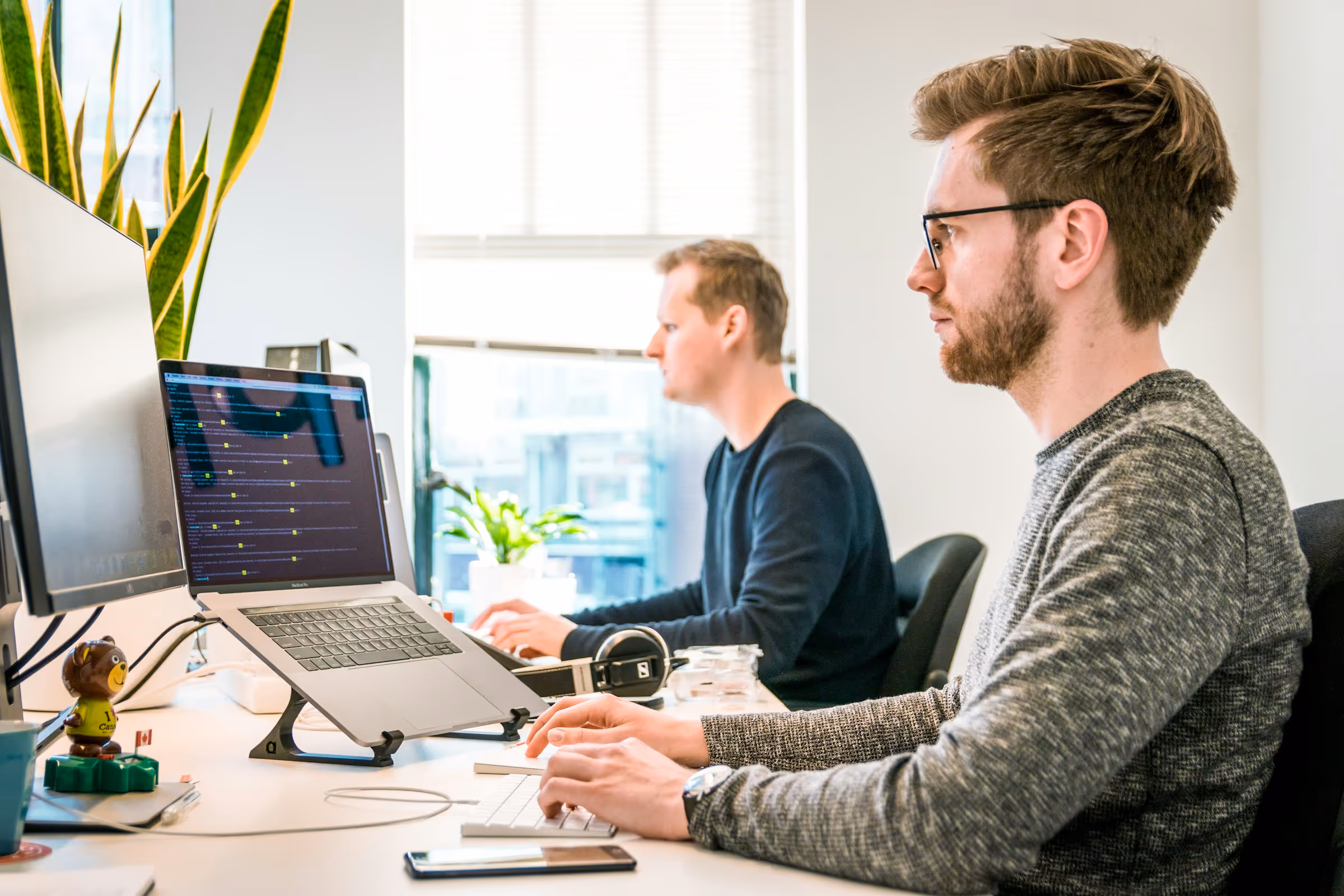 Two men working focused at desks with laptops and monitors in a bright office space with plants.