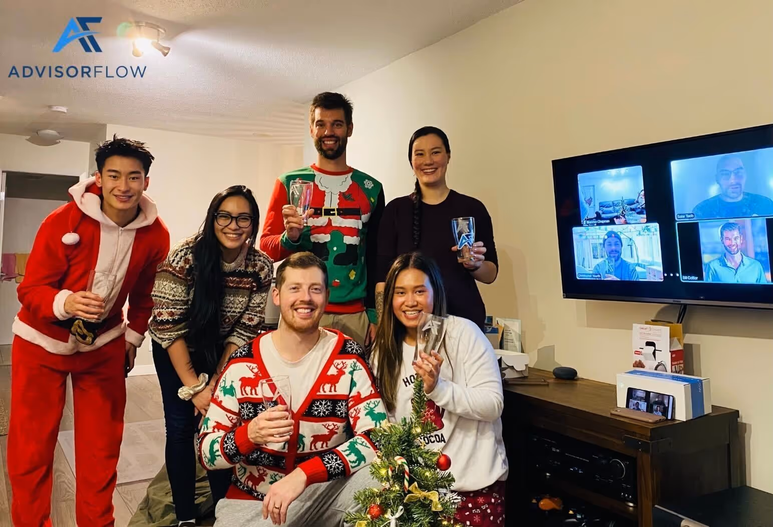 Six people dressed in festive holiday attire smiling and holding glasses, with a small decorated Christmas tree in front, and four others visible on a video call screen in the background.