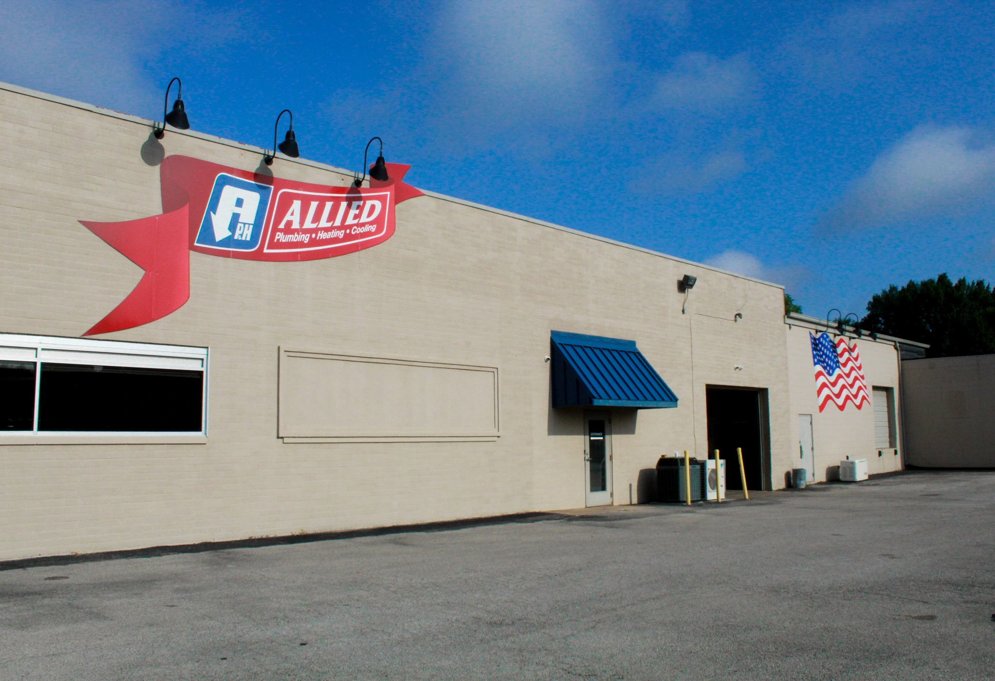A beige industrial building with a sign reading "Allied Plumbing, Heating, Cooling," a blue awning, and an American flag mural.