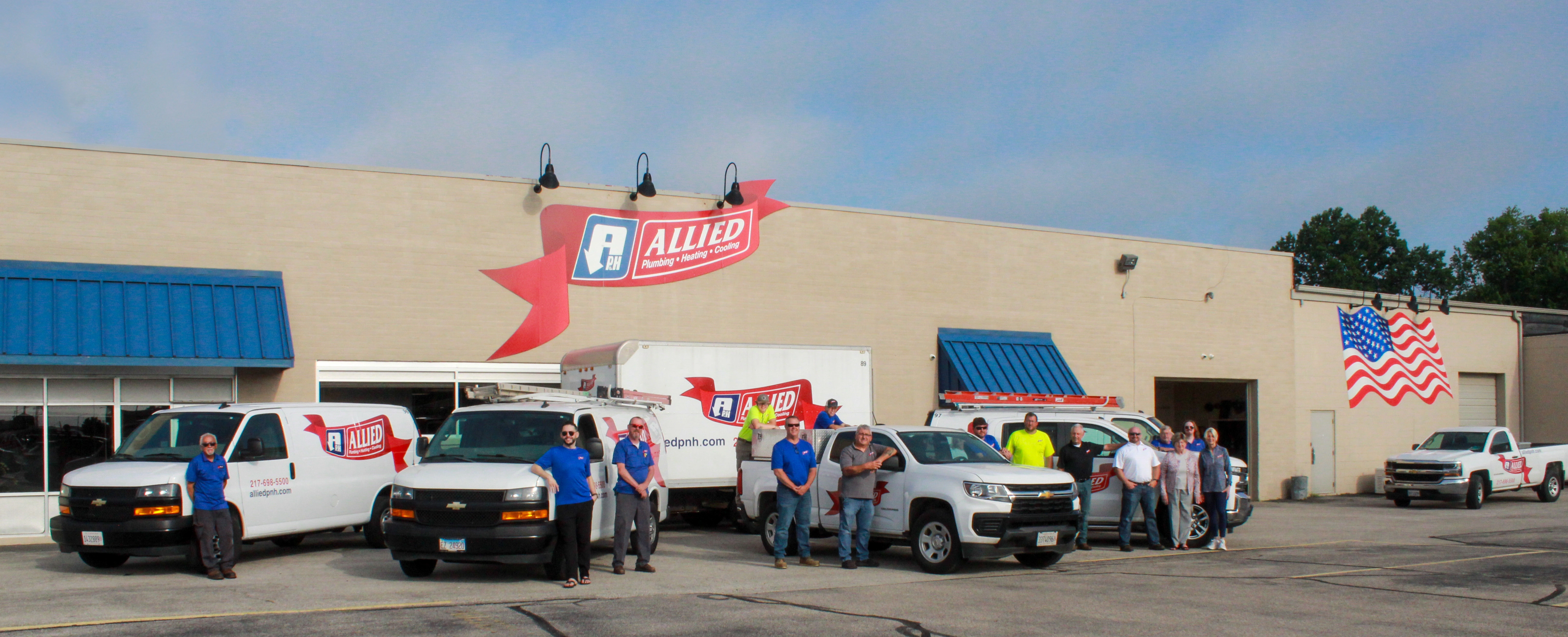 Group of people in casual attire stand in front of a building with blue awnings, Allied logo, and multiple branded vans.
