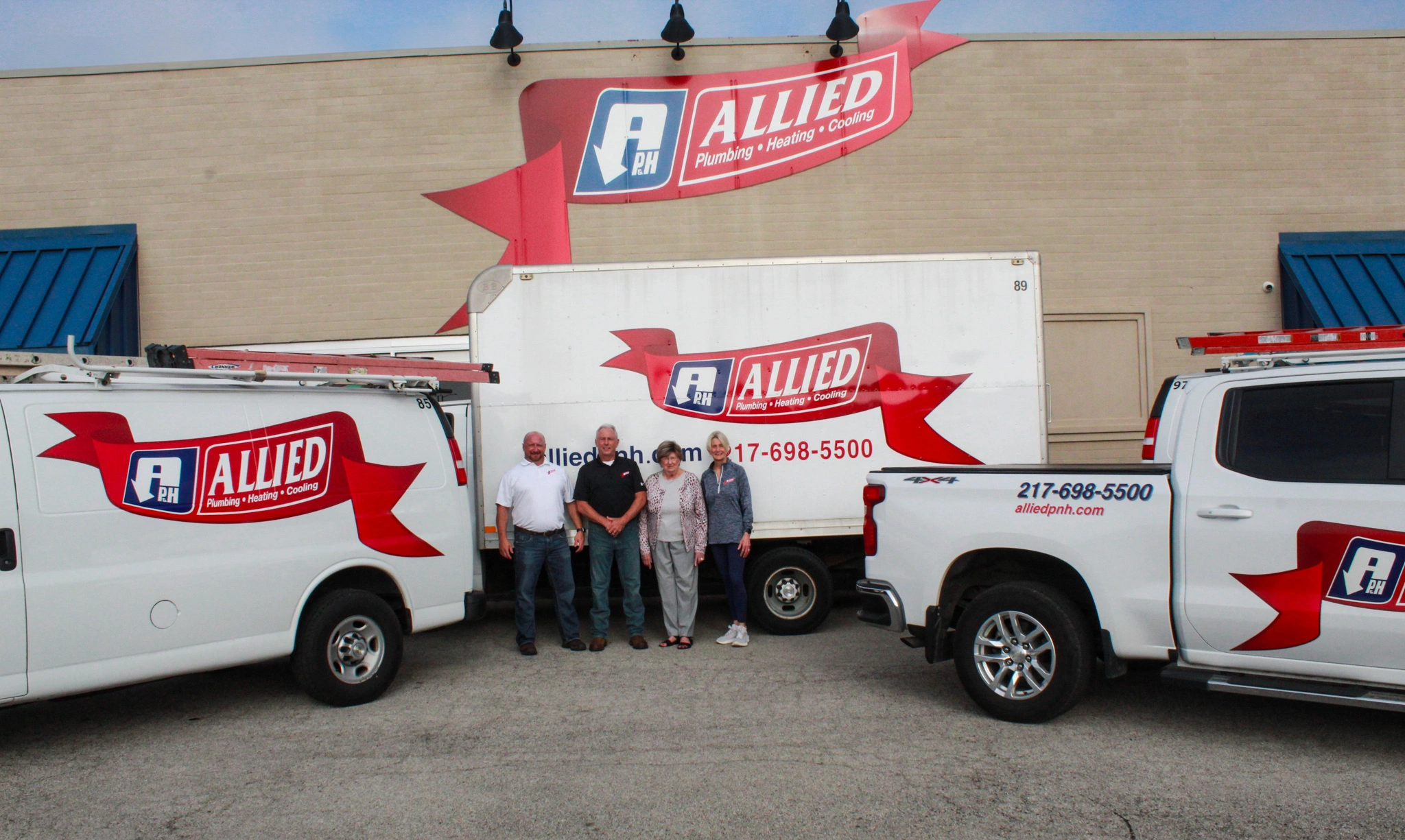 Four people stand in front of Allied Plumbing trucks, parked by a building with Allied signage.