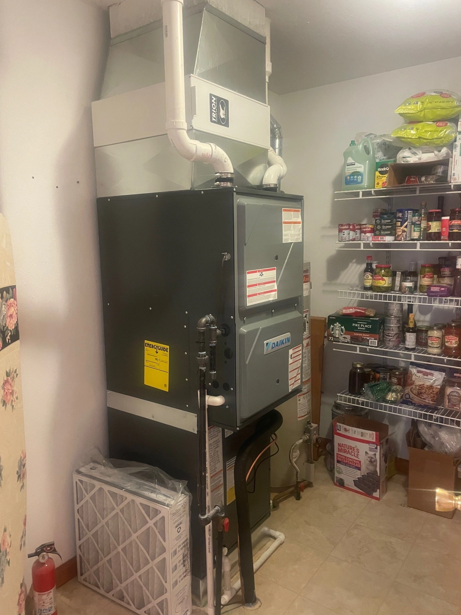 A large gray furnace with pipes is in a utility room. Shelves on the right are stocked with food and household items.