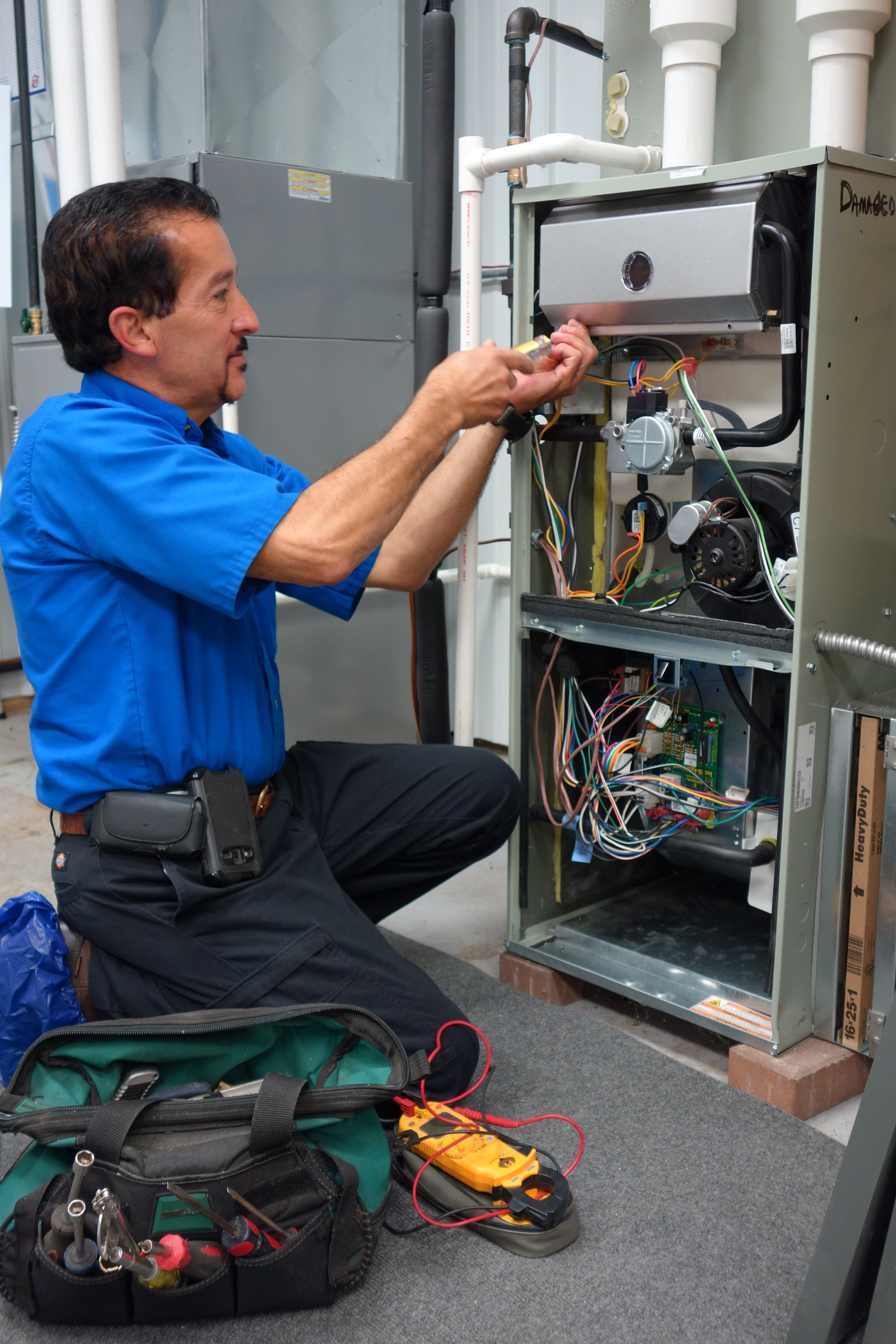 A technician in a blue shirt kneels on the floor, repairing an HVAC unit with multiple wires exposed.