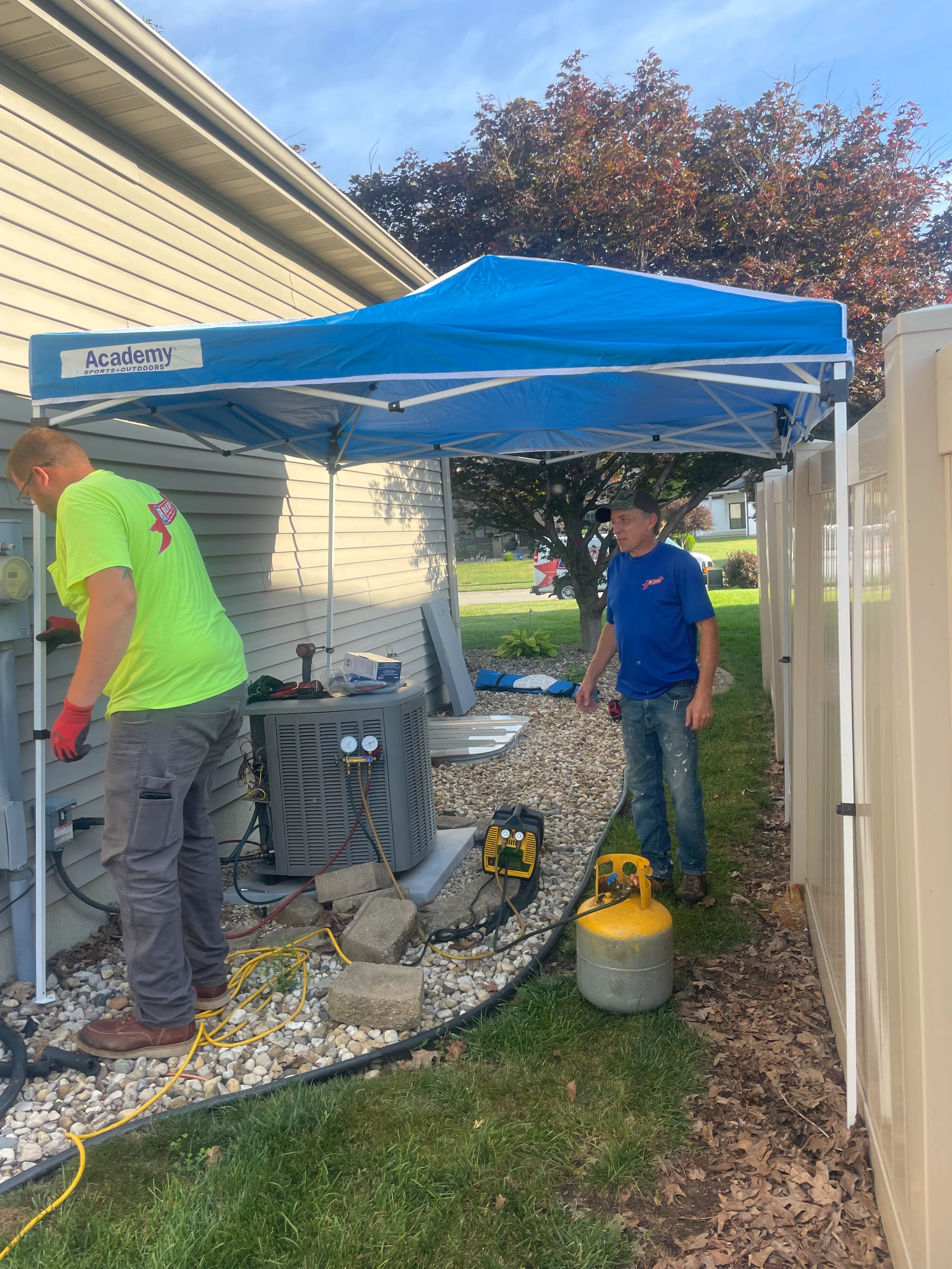 Two workers set up air conditioning equipment under a blue canopy beside a house.