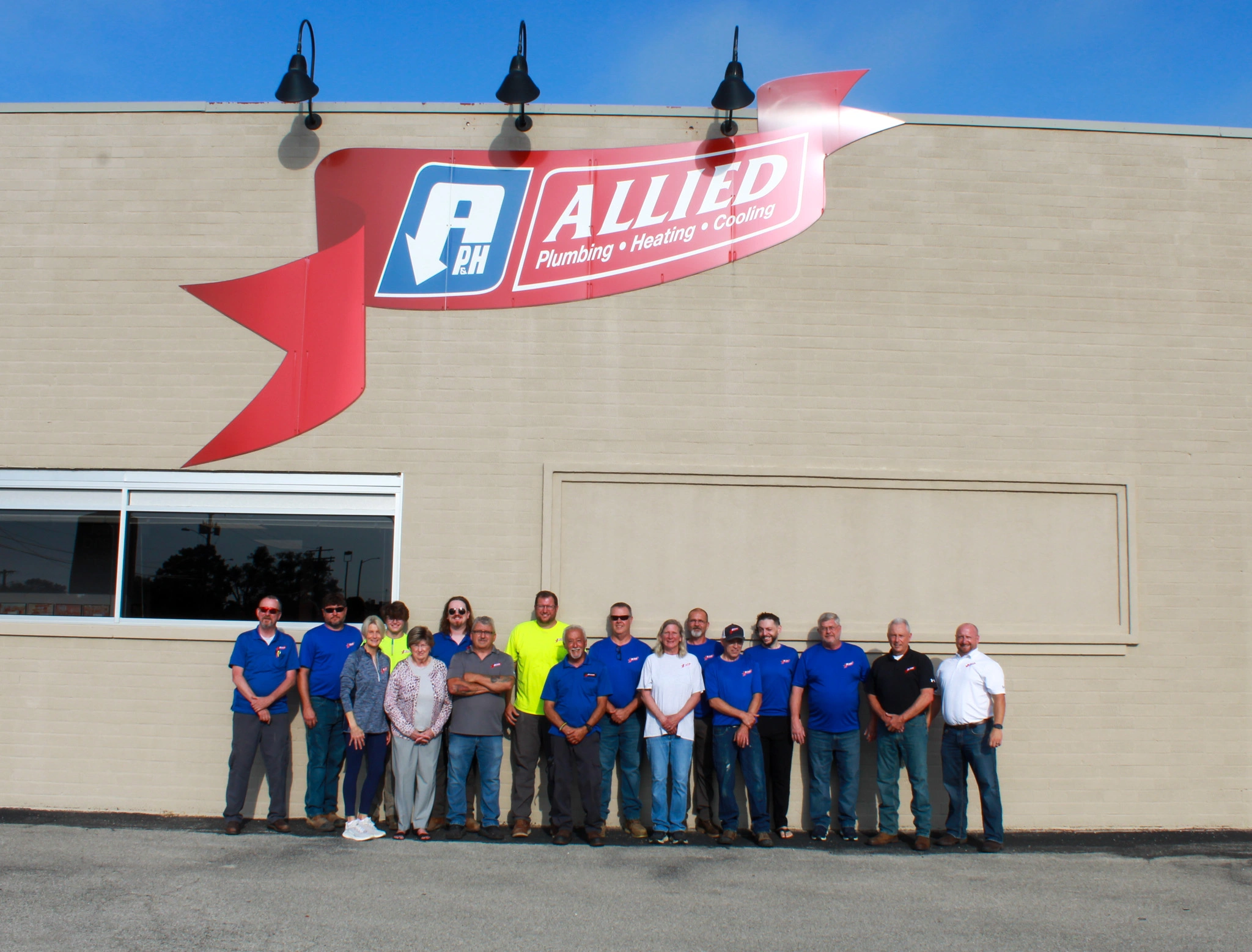 A group of fifteen people stands smiling in front of a beige building. A large red sign above reads "Allied Plumbing Heating Cooling," conveying a team spirit.