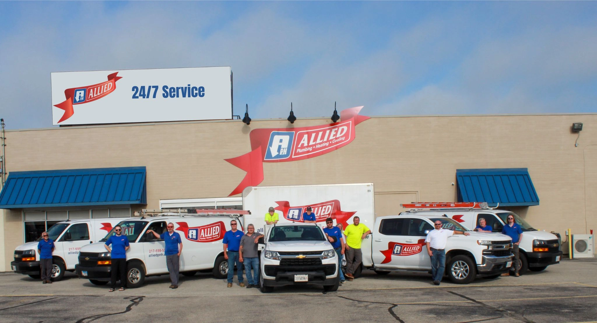 A group of workers in blue and neon shirts stand in front of company vans and a truck with Allied logos.