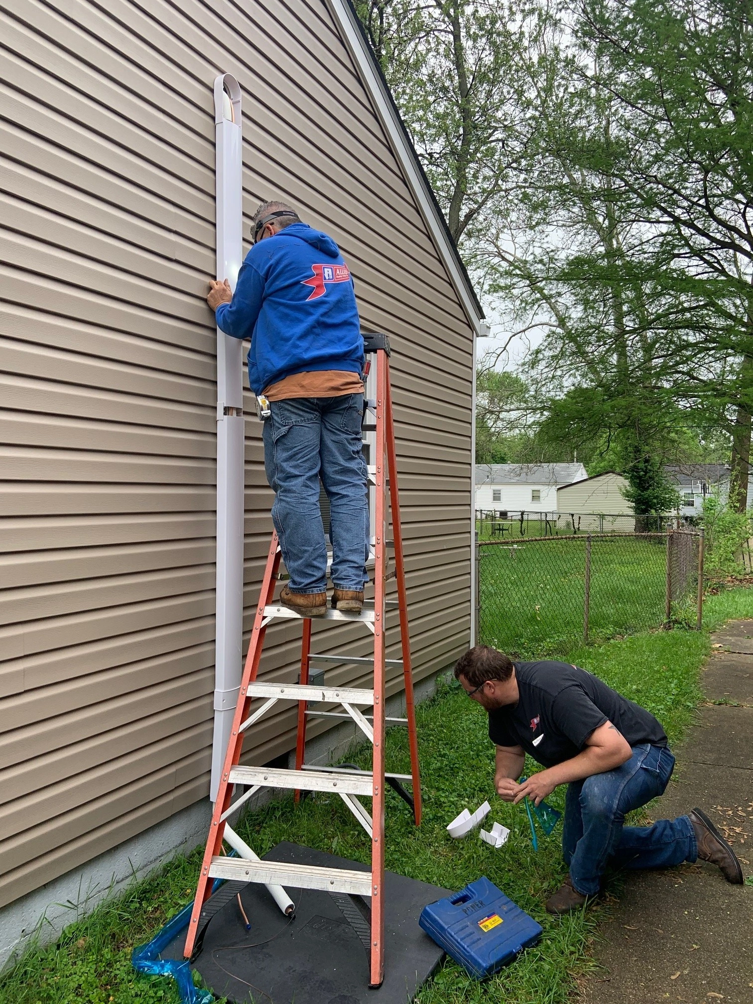 Two men are working on a beige house exterior. One stands on a ladder, wearing a blue hoodie, adjusting a vertical panel.