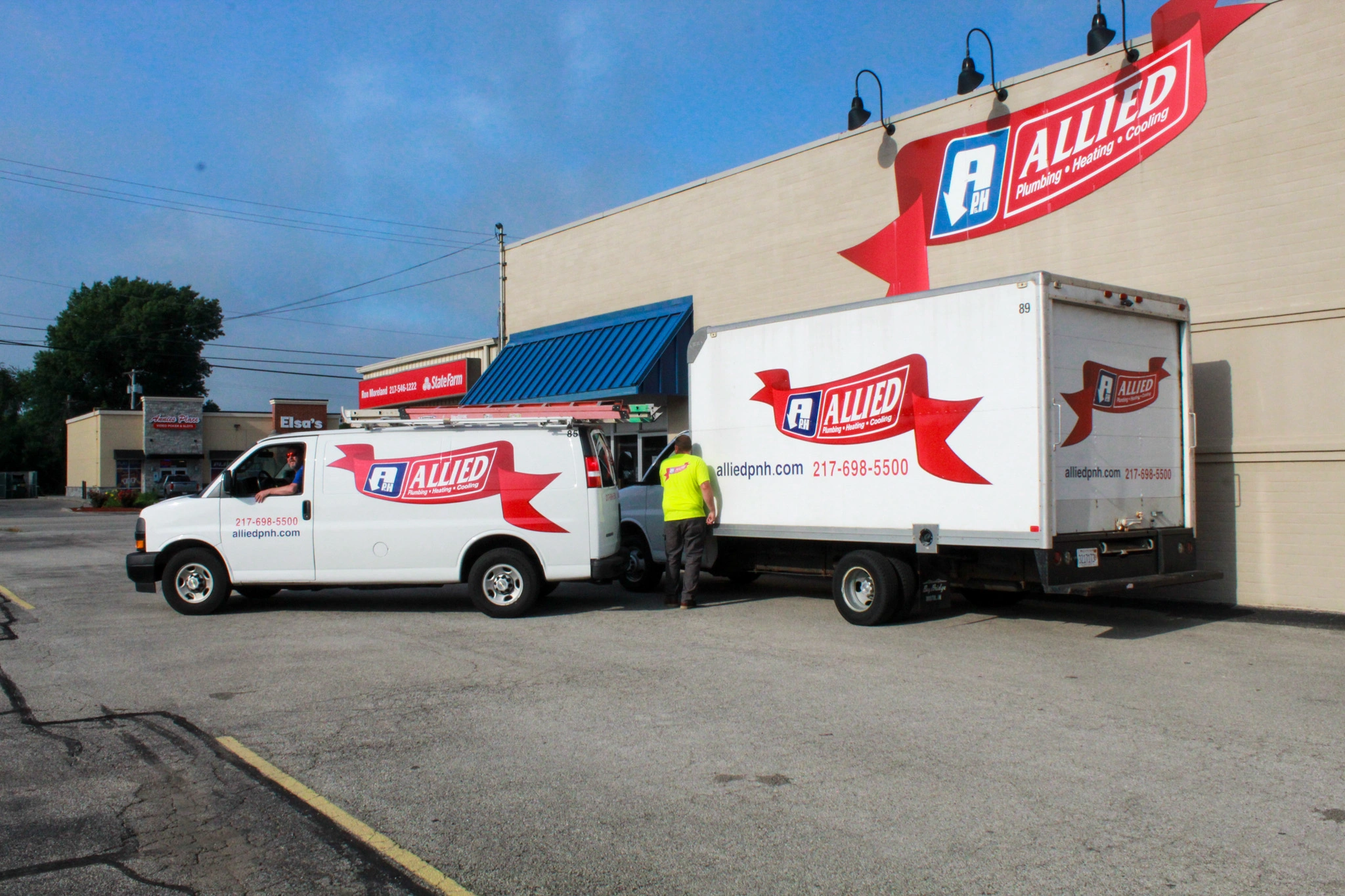 Two white Allied Plumbing trucks are parked outside a beige building with a blue awning. 