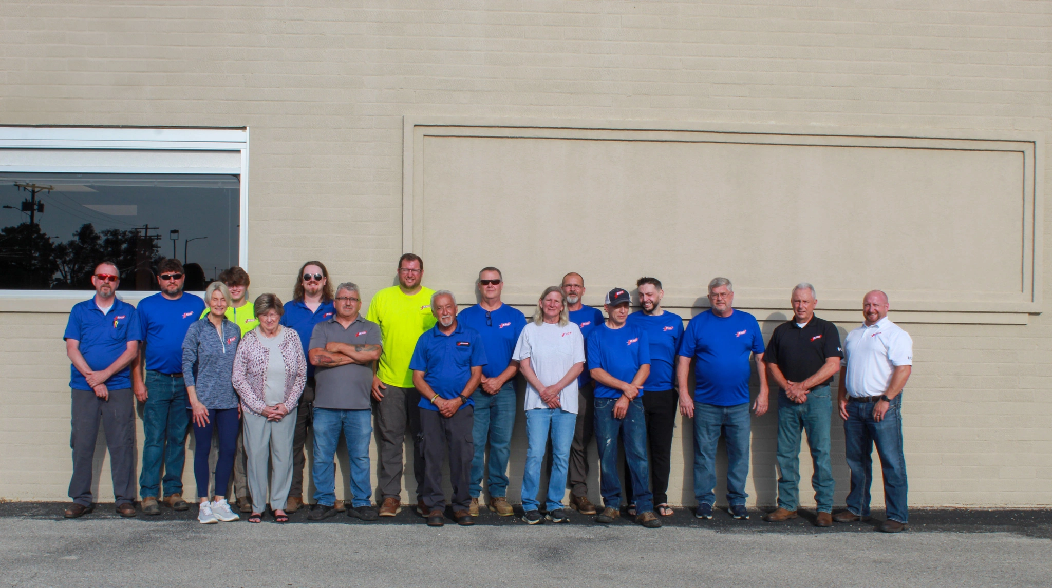 A group of 18 people stand side by side against a beige brick wall. Most wear blue shirts