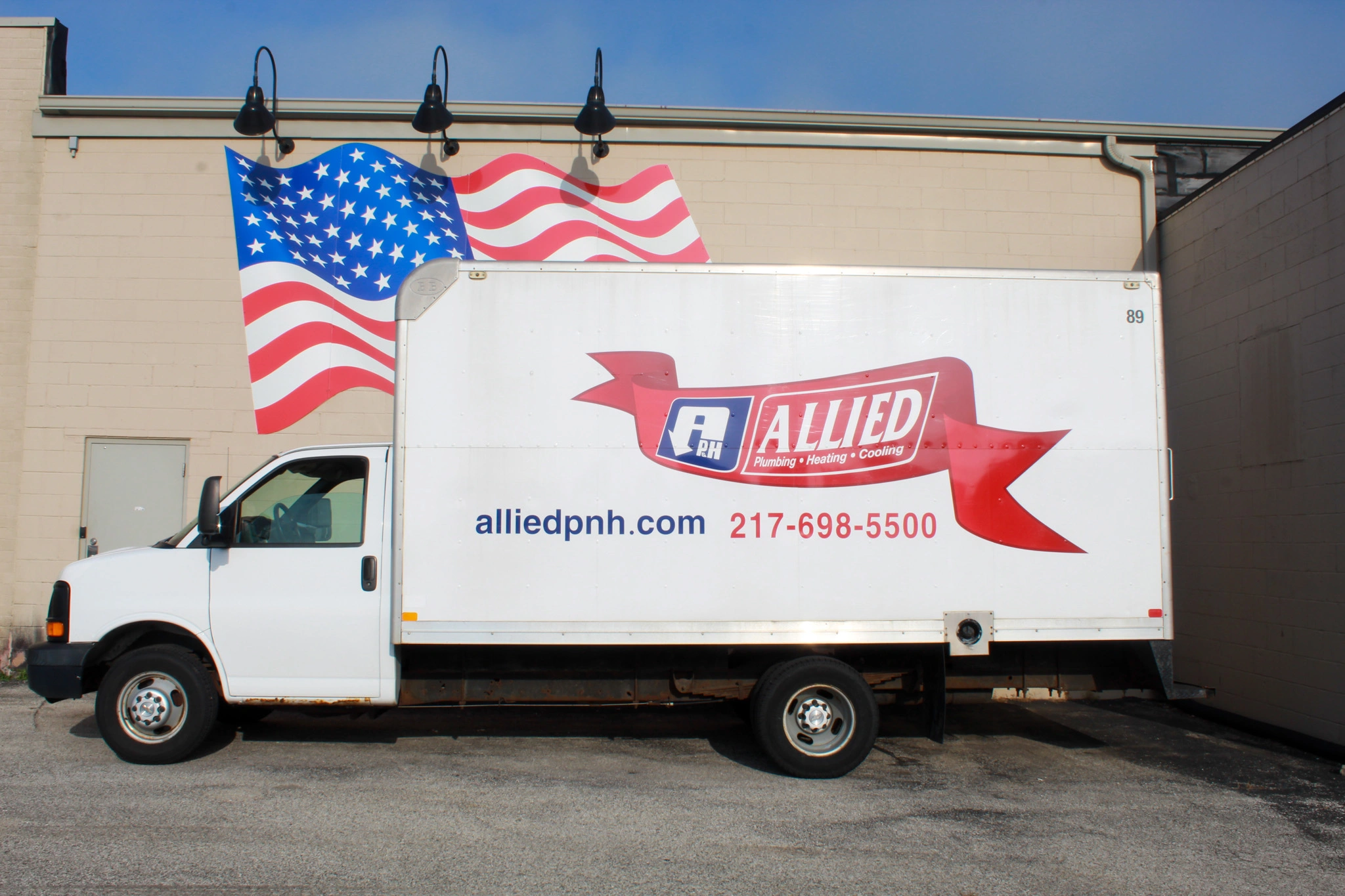A white delivery truck is parked beside a beige wall painted with a large, waving American flag. The truck displays "Allied" logo, website, and phone number.