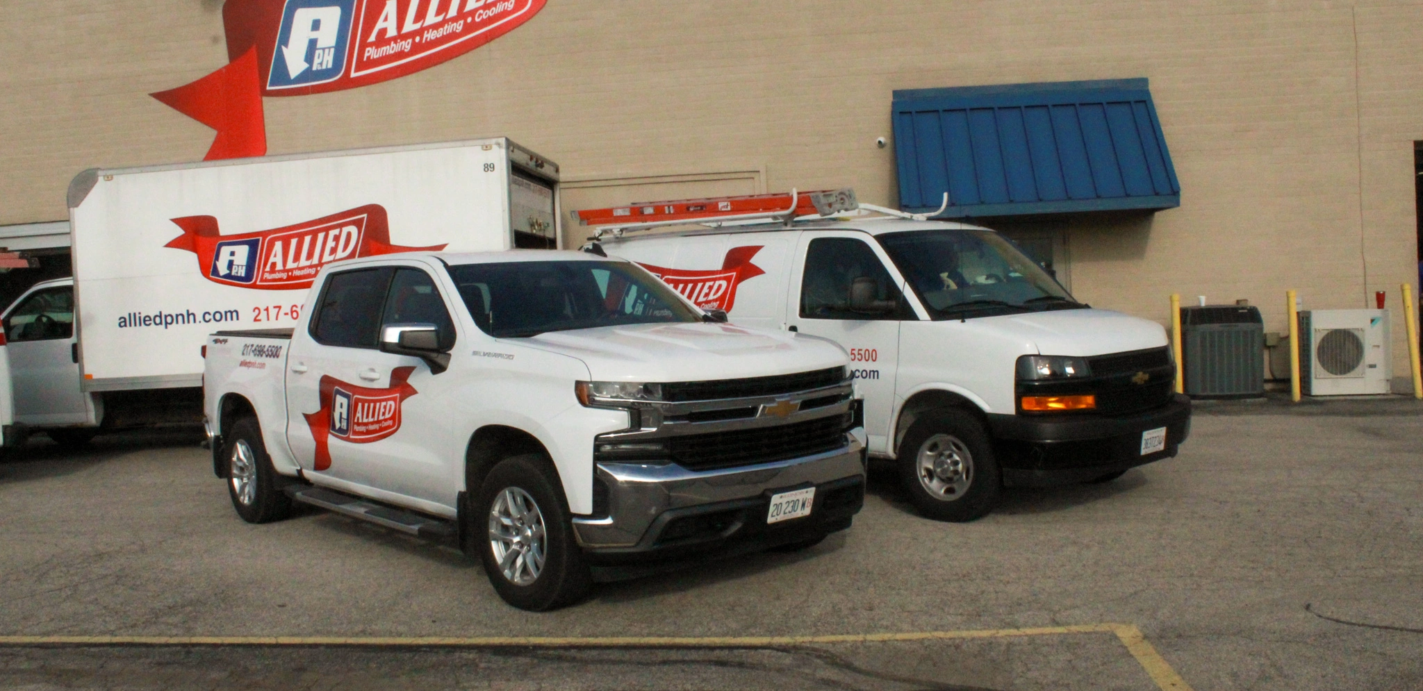 Parking lot scene with two white Allied Plumbing vehicles; a truck and a van, parked side by side.