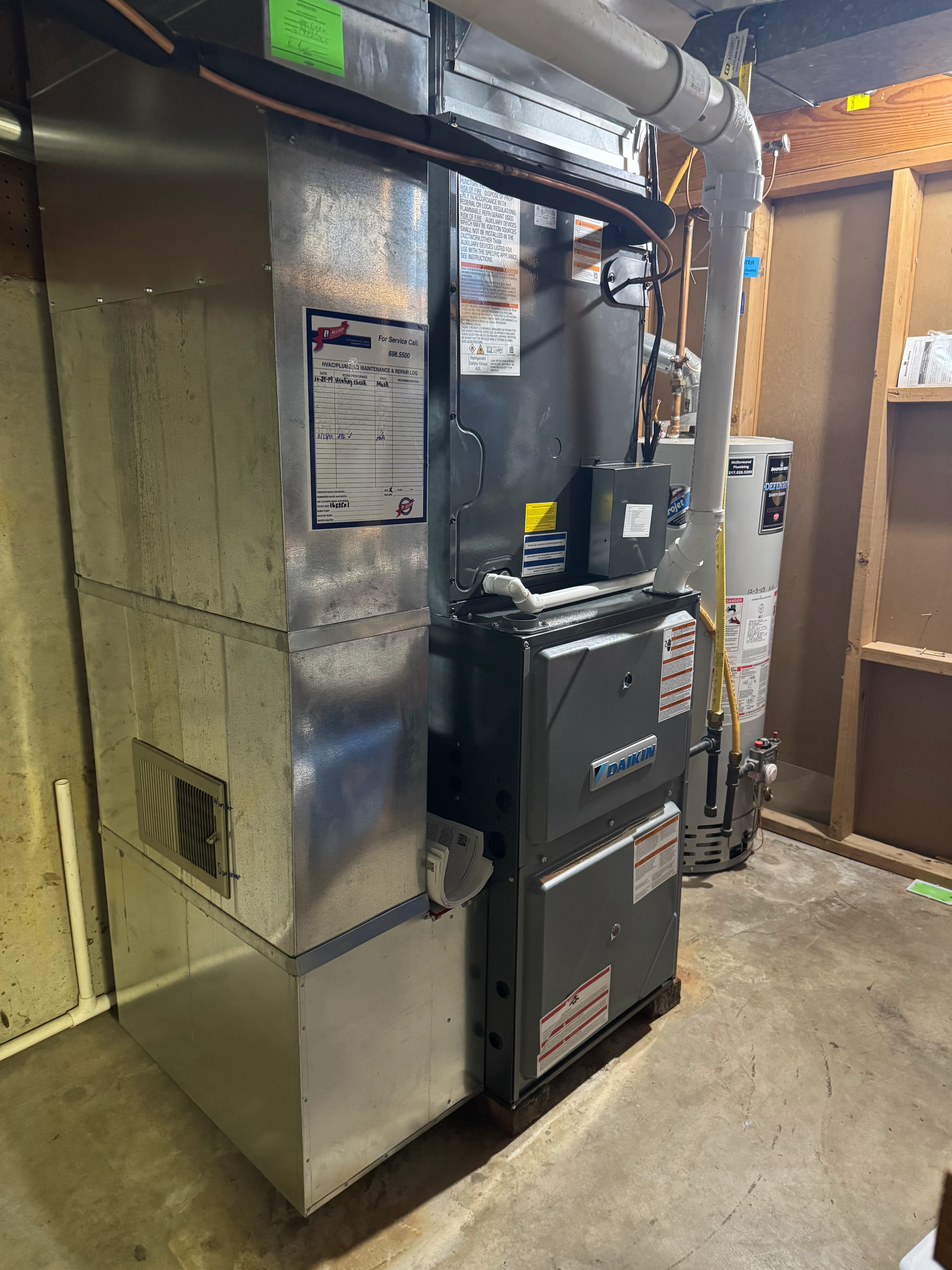 A large metal furnace and water heater are situated in a basement with concrete floors.