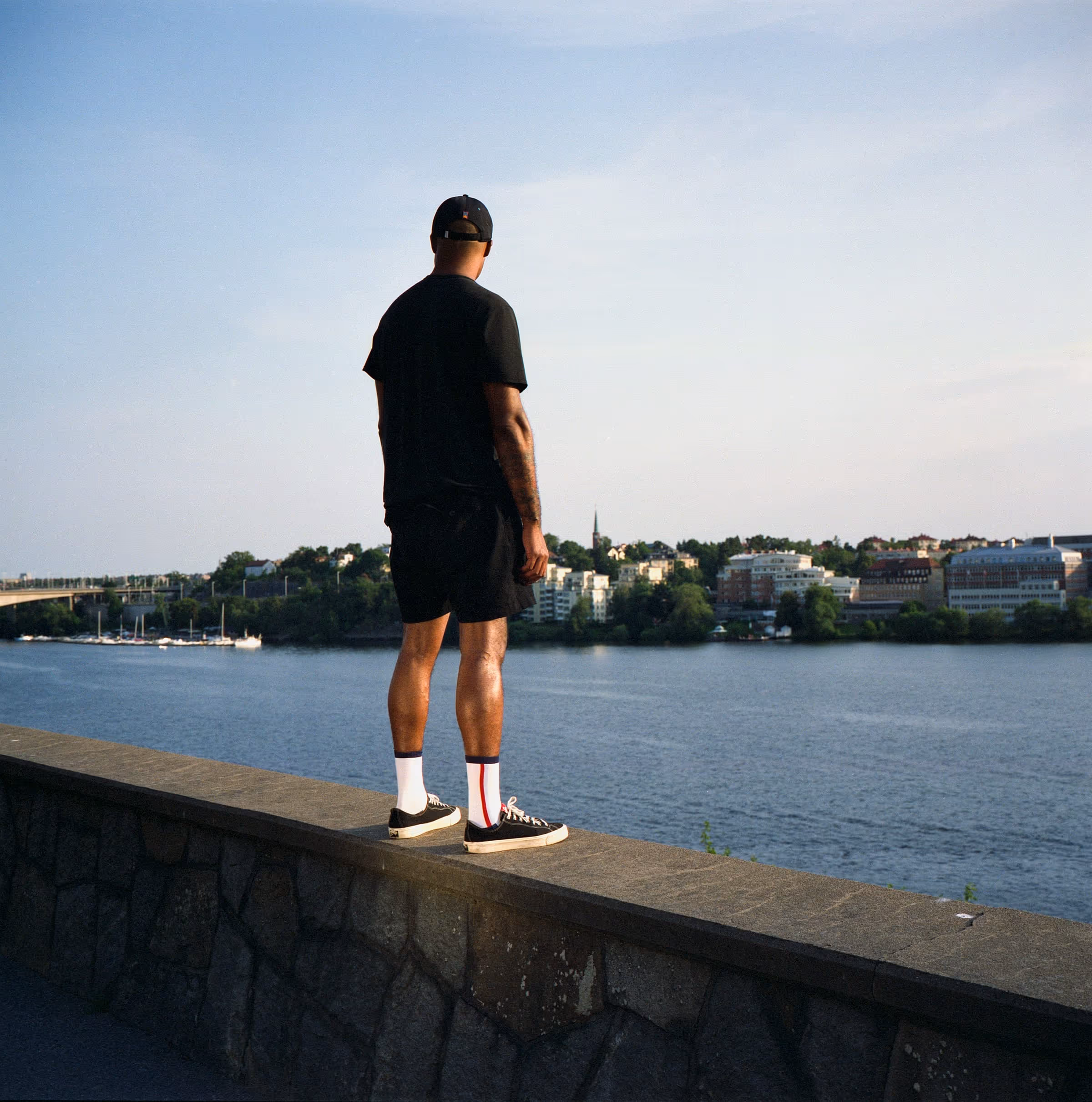 Man dressed in black standing on a stone wall overlooking a calm river with buildings and a bridge in the background under a clear sky.
