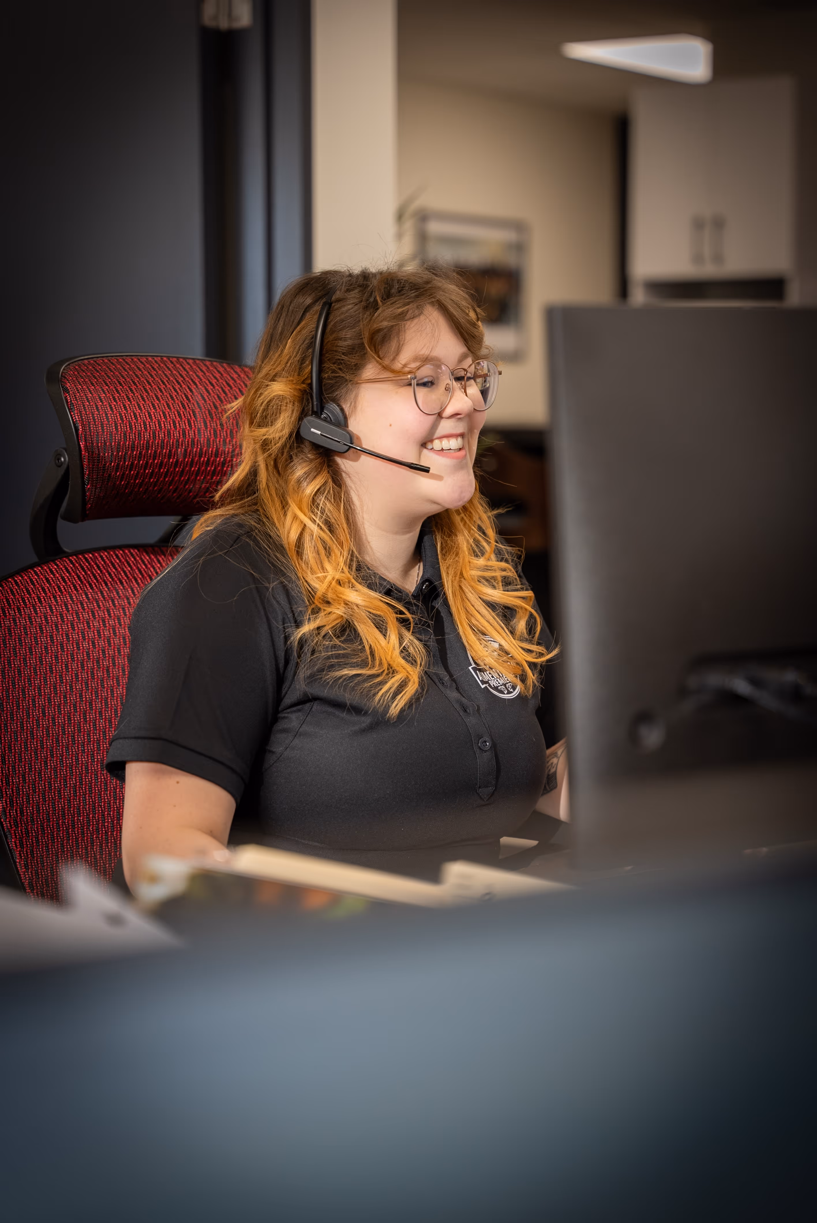 Jeune femme souriante avec casque et lunettes travaillant devant un ordinateur dans un bureau.