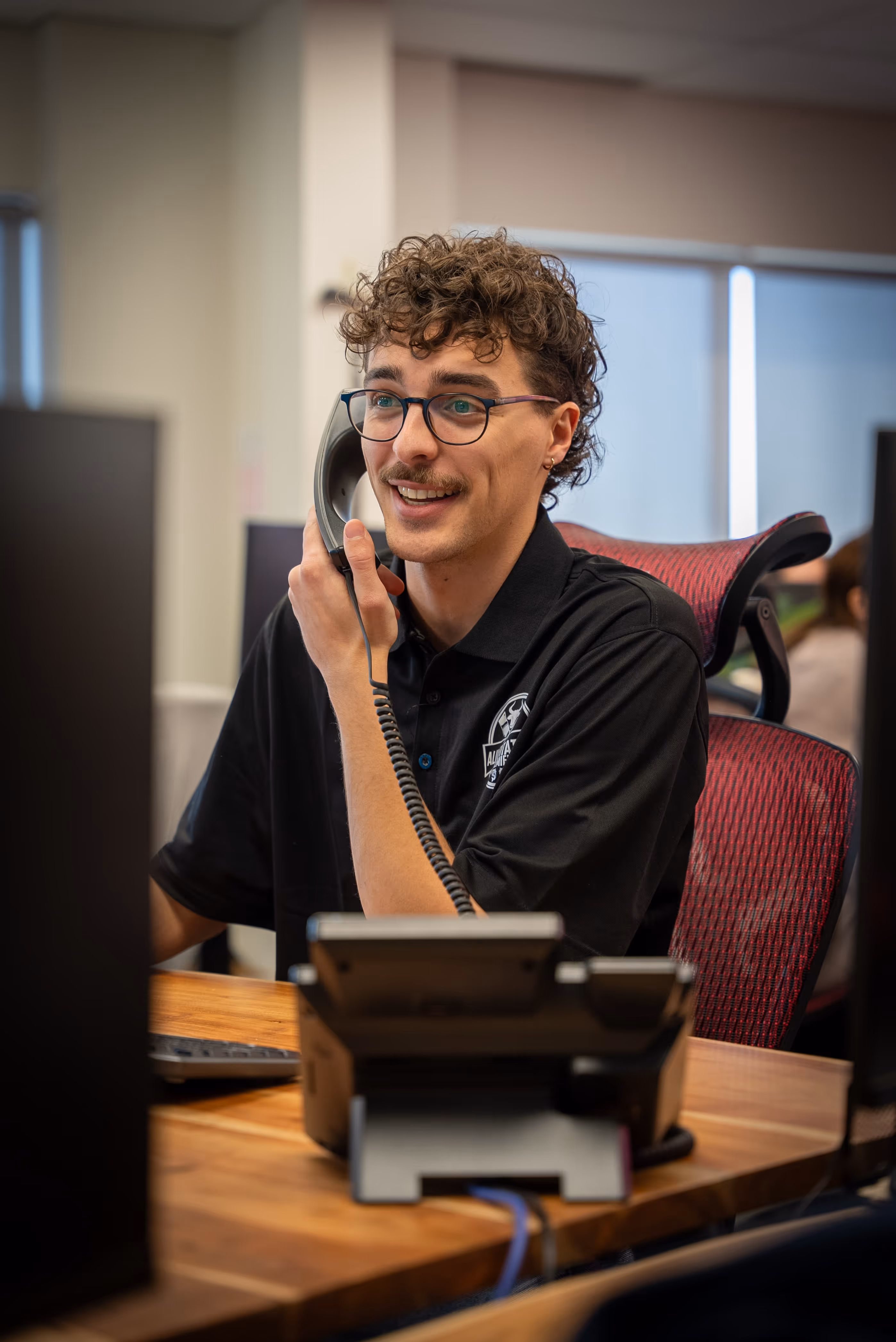 Un homme souriant portant des lunettes et un polo noir parle au téléphone dans un bureau.