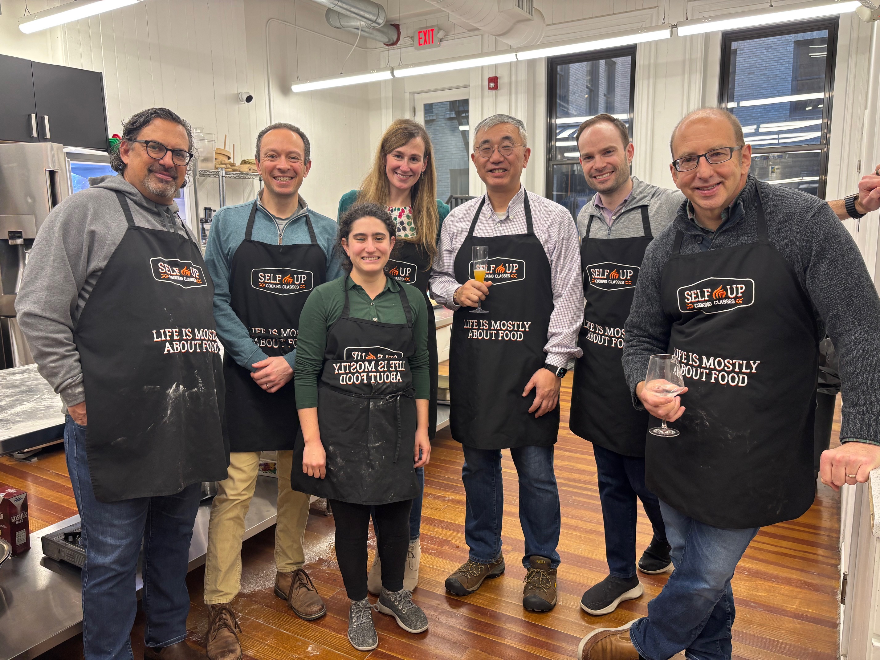 Group of seven GreenieRE team members in aprons reading 'Life is mostly about food' posing in a bright kitchen, some holding glasses.