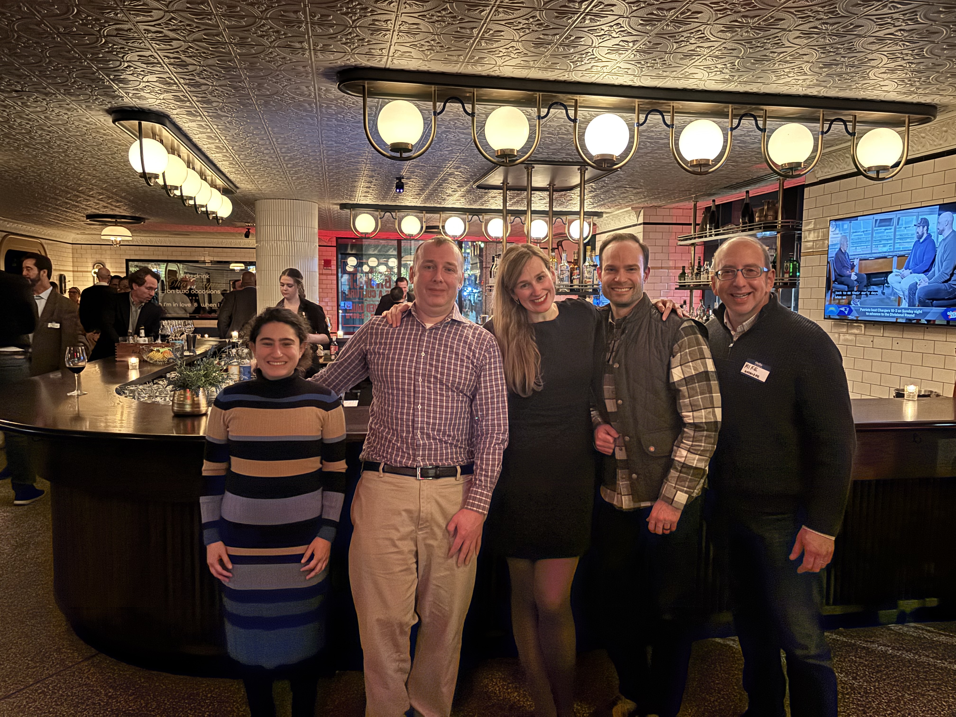 Five GreenieRE team members smiling and posing together inside a warmly lit bar with round ceiling lights and a TV on the wall.