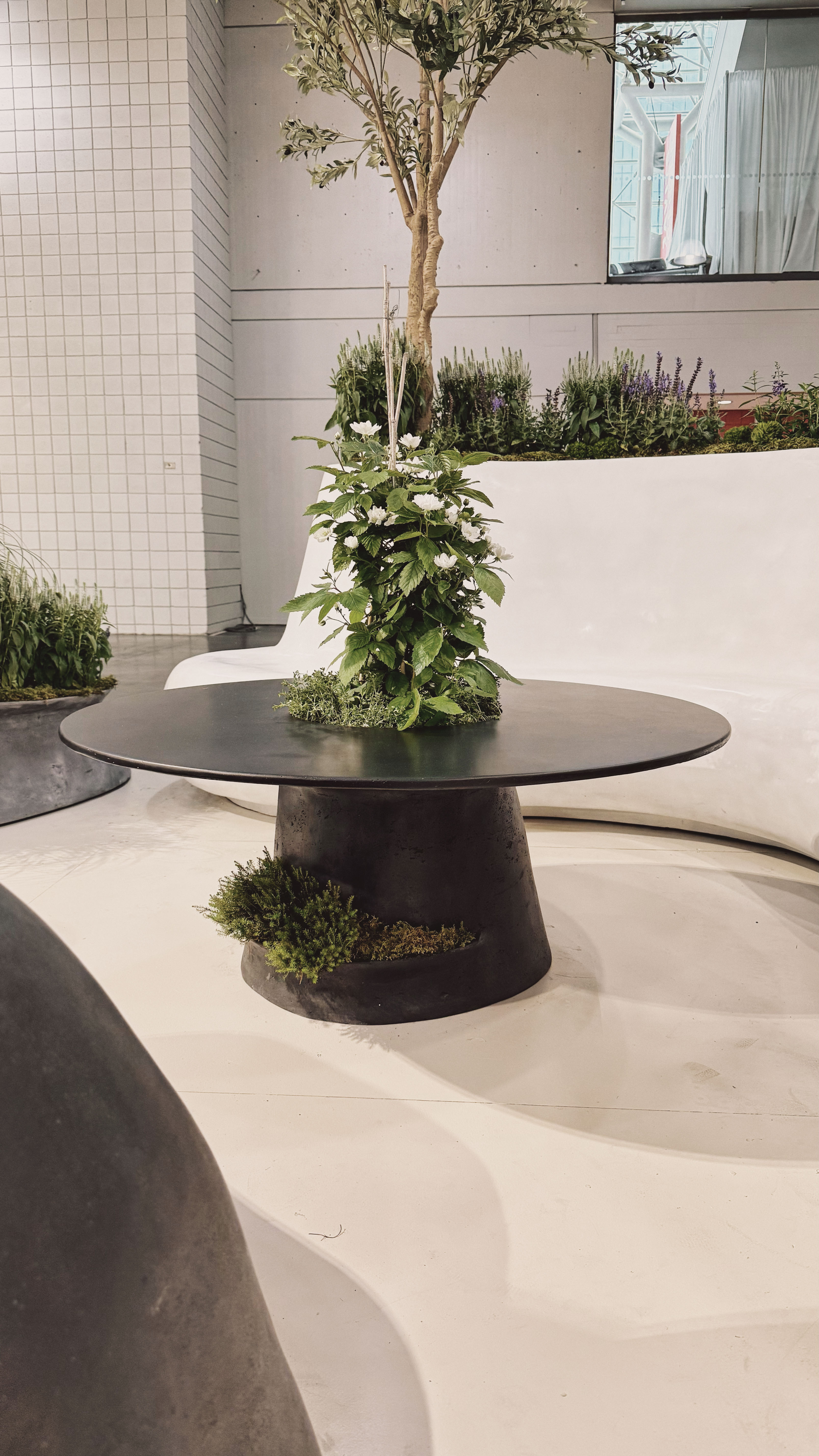 A black concrete table with a planter built into the base and the center of the table top. A large plant sprouts out of the tabletop while a smaller plant grows out of the base.