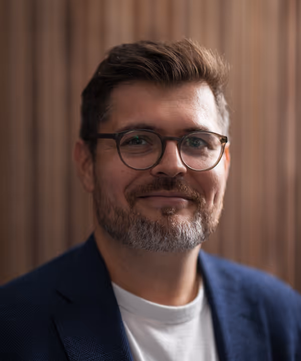 Man with glasses and beard smiling, wearing blue blazer and white t-shirt, short brown hair.