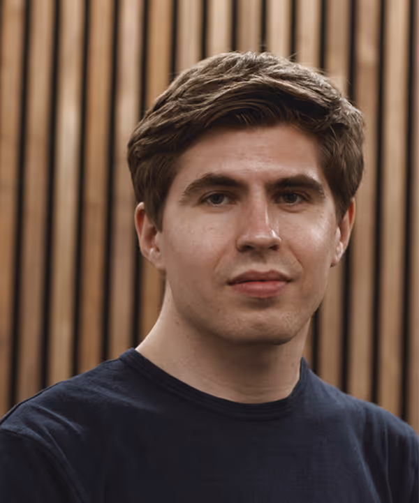Young man with short brown hair wearing a black t-shirt standing in front of wood slats.