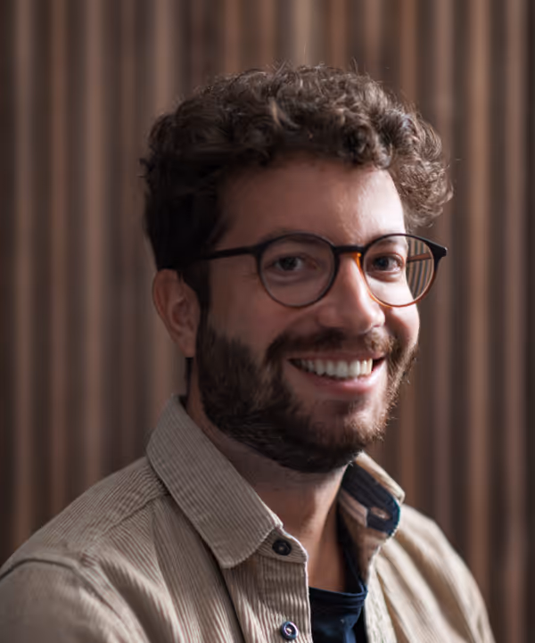 Man with curly hair and beard smiling, wearing glasses and a tan button-up shirt indoors.