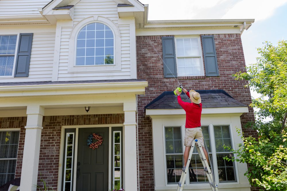 Person in red shirt on ladder spraying insecticide on upper wall of brick and siding house.
