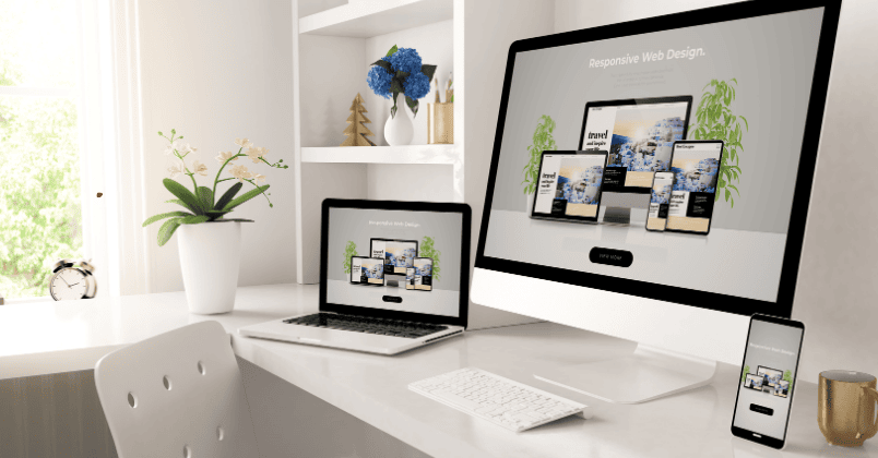 Modern white desk setup with an iMac, laptop, and smartphone displaying responsive web design pages, with potted plants and a gold cup nearby.
