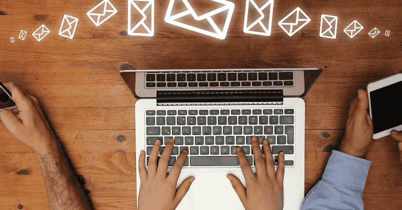 Top view of hands working on a laptop keyboard with two other hands holding smartphones on each side and floating email icons above the laptop.