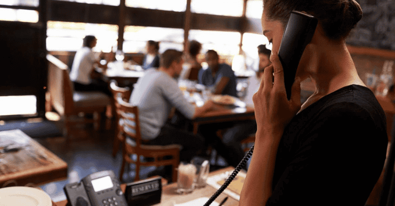 Woman speaking on a telephone in a busy restaurant with patrons dining in the background.