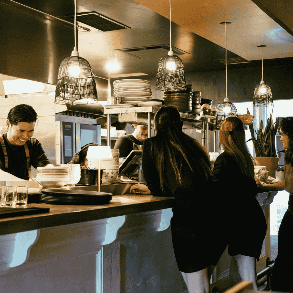 People interacting behind a restaurant counter under hanging lamps with stacks of plates and bowls on shelves.