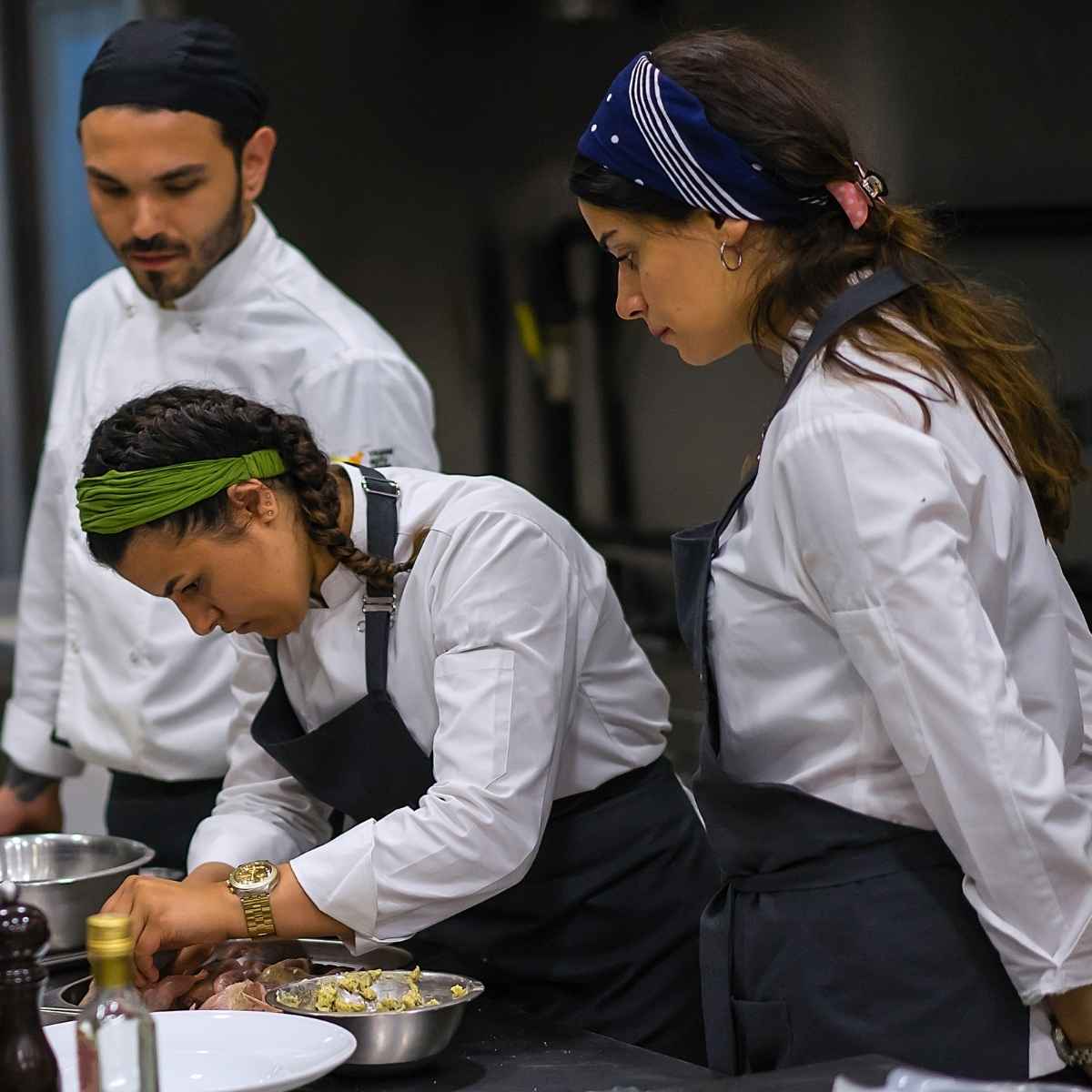 Three chefs in white uniforms and dark aprons focused on preparing food in a professional kitchen.