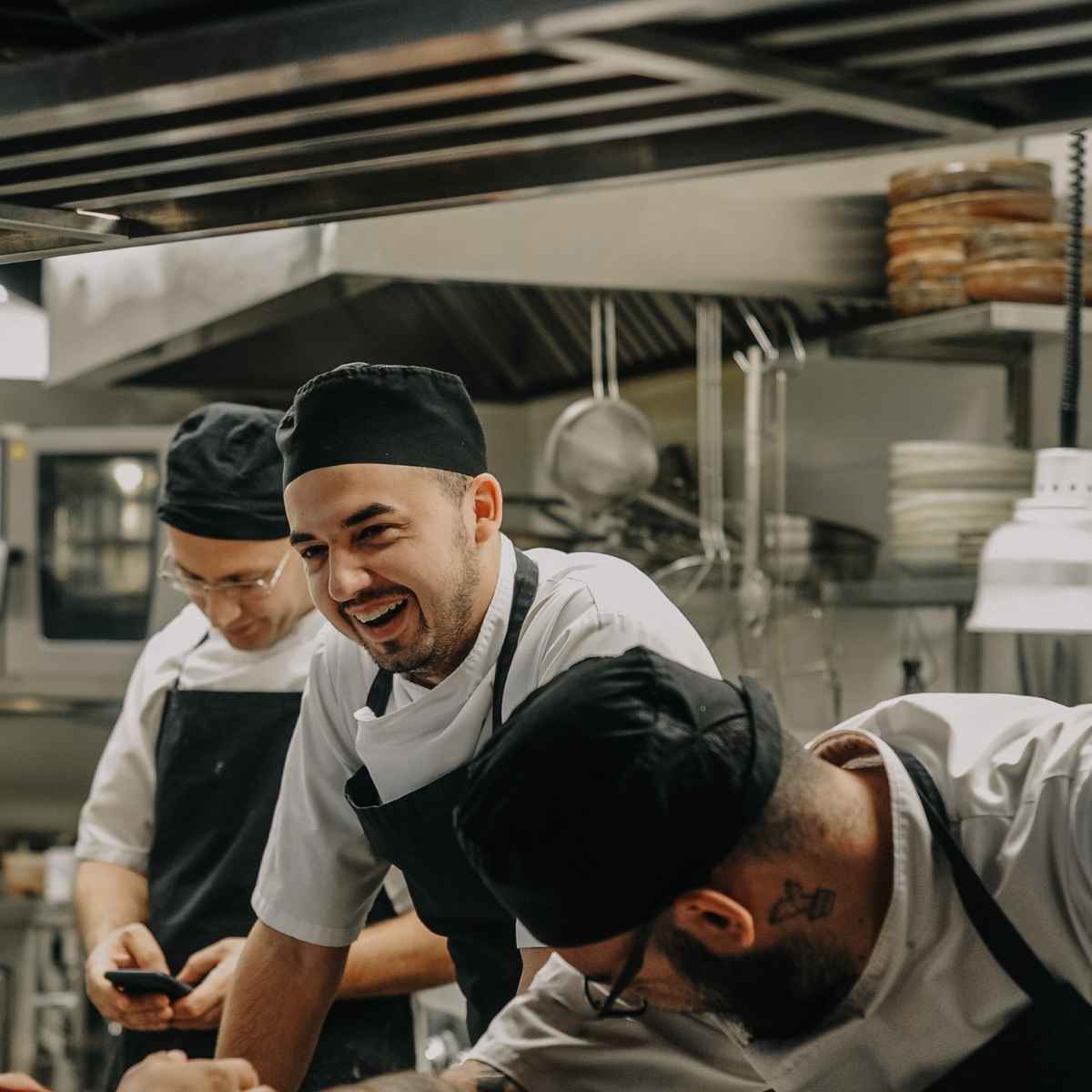 Three chefs in white uniforms and black aprons and hats working and smiling in a commercial kitchen.